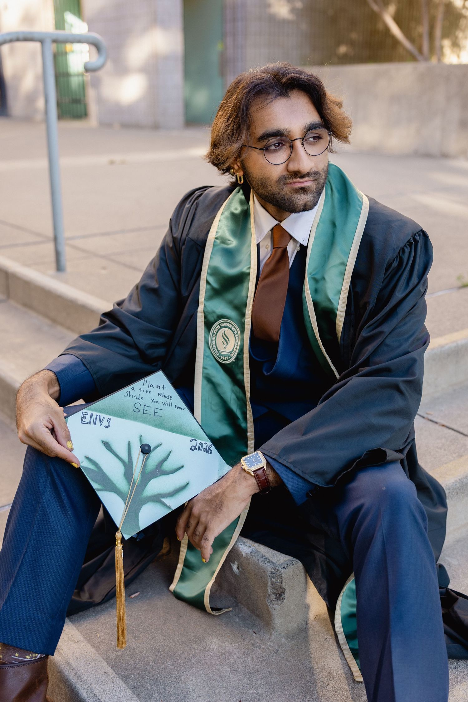 Graduate in black gown and green stole sits holding decorated cap outdoors.