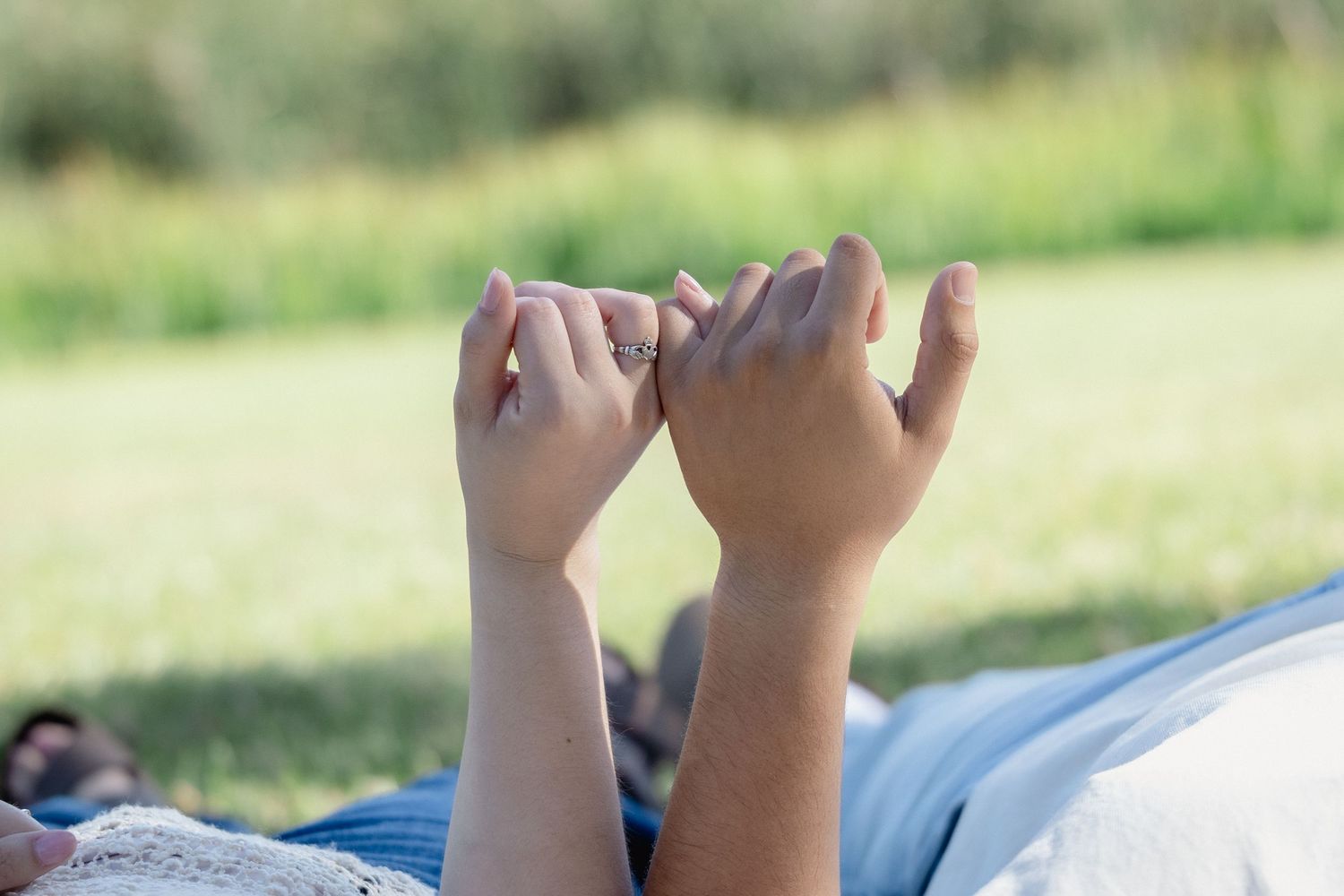 Couple holding pinky fingers together outdoors on a sunny day with green grass in the background.