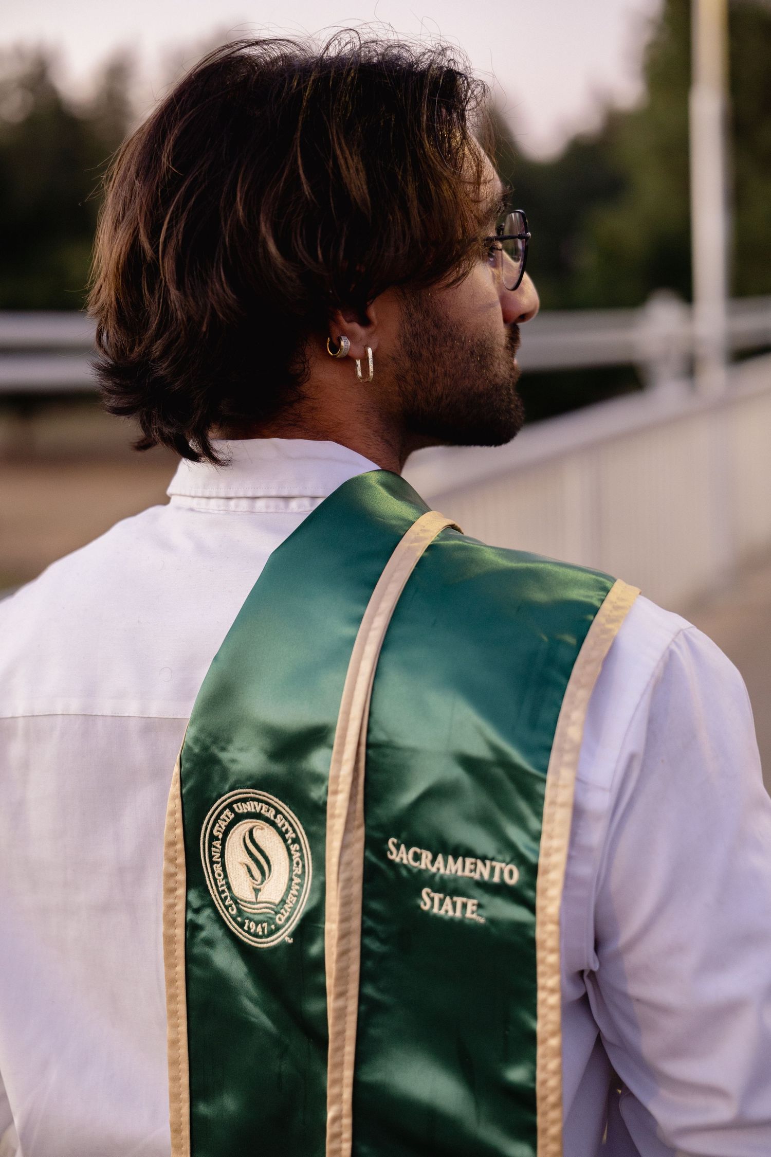 Sacramento State University graduate wearing green and gold sash over white dress shirt at outdoor graduation ceremony.