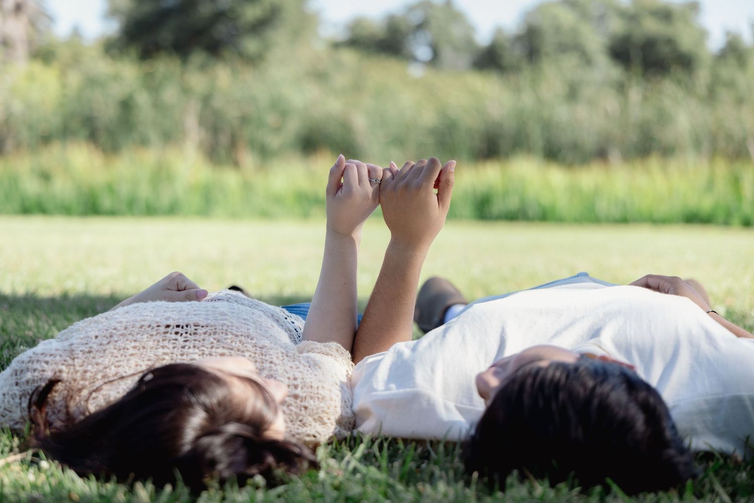 Couple lying on green grass in a park, holding feet up together on a sunny day.