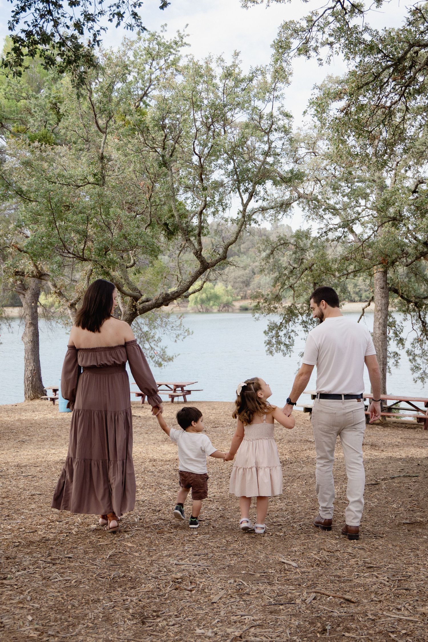 Family of four holding hands walking toward a serene lake surrounded by trees in a park setting.