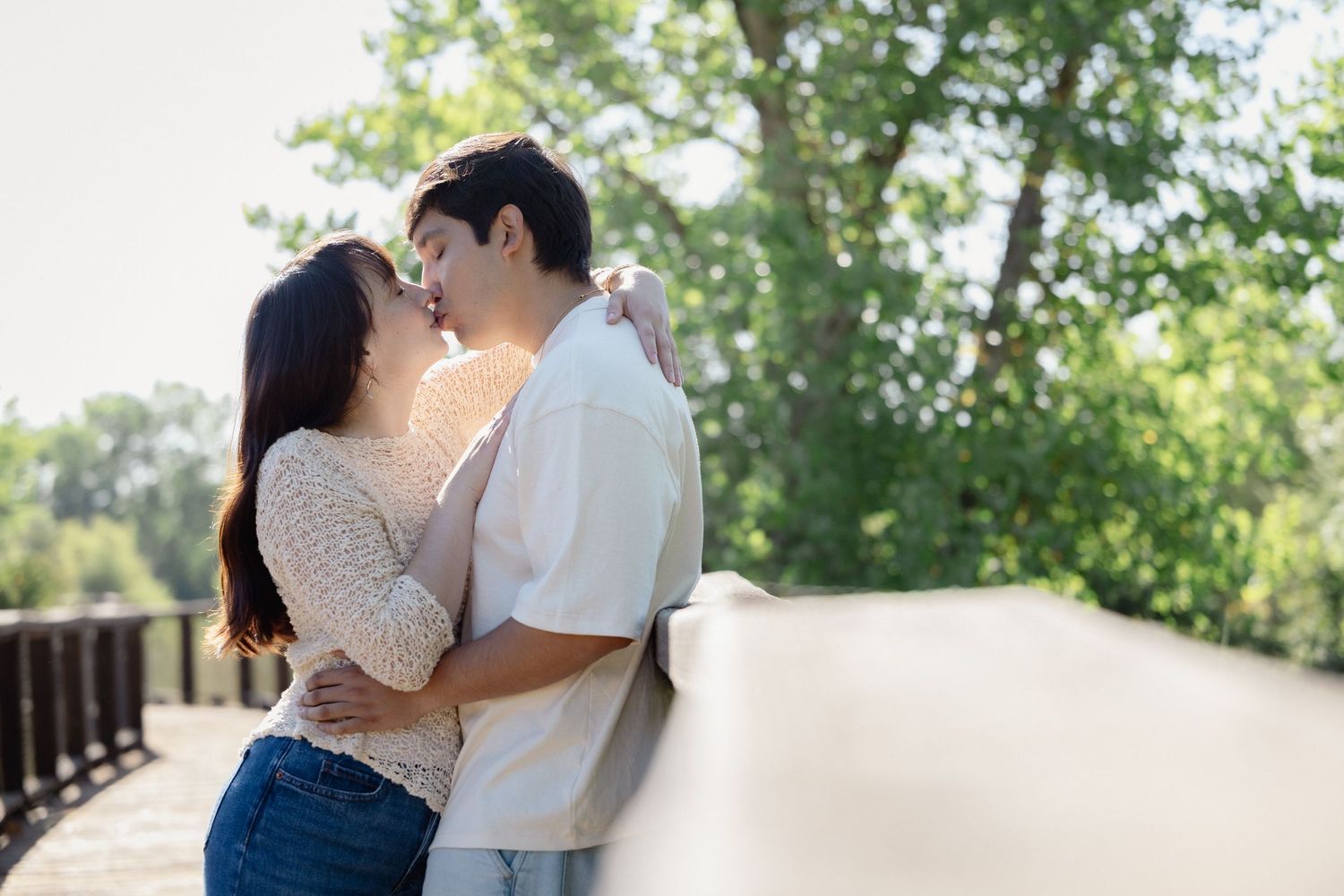 Couple sharing a romantic kiss on a bridge surrounded by lush green trees on a sunny day.
