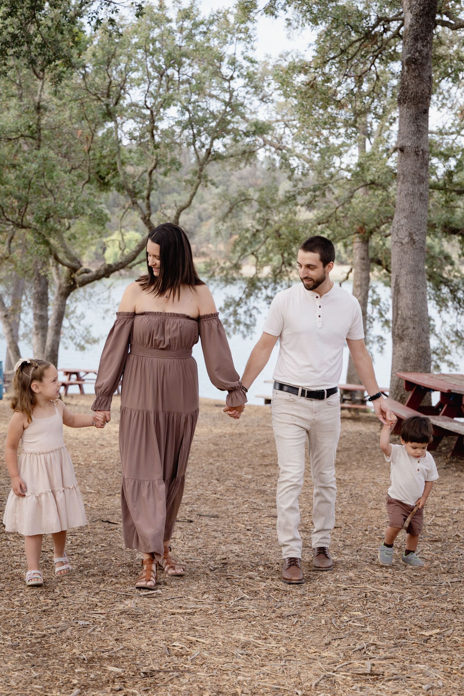 A happy family of four walks hand-in-hand through a tree-lined park near a lake on a sunny day.