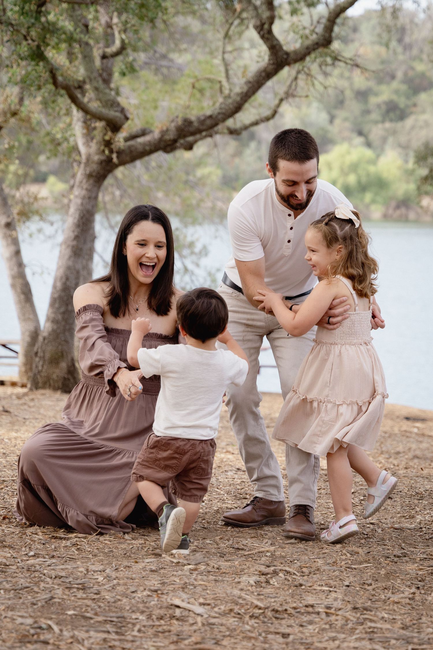 Happy family of four playing outdoors near a lake, parents laughing with toddler boy and young girl in nature.