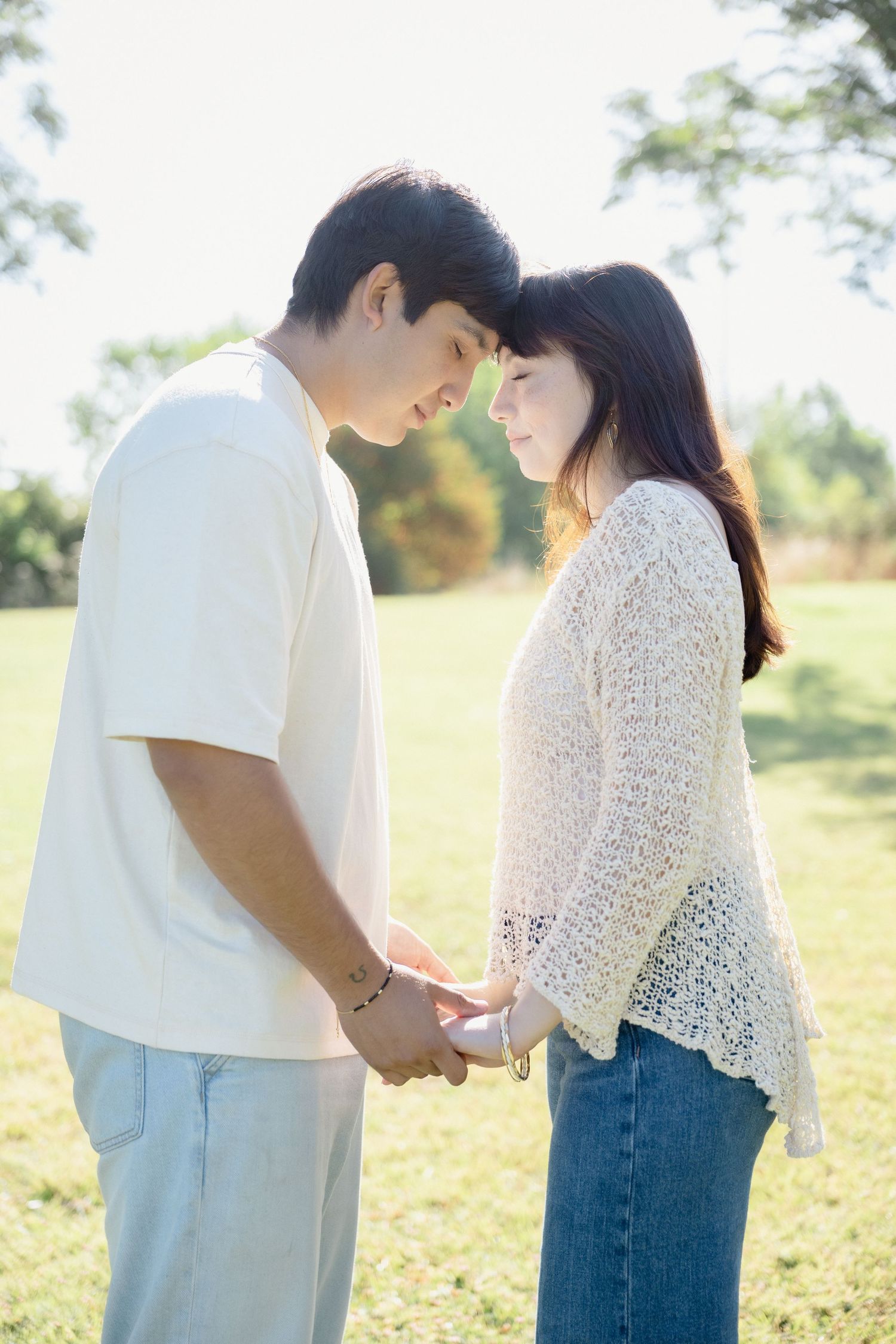 Young couple holding hands and touching foreheads in a sunny park, wearing casual white tops and jeans.