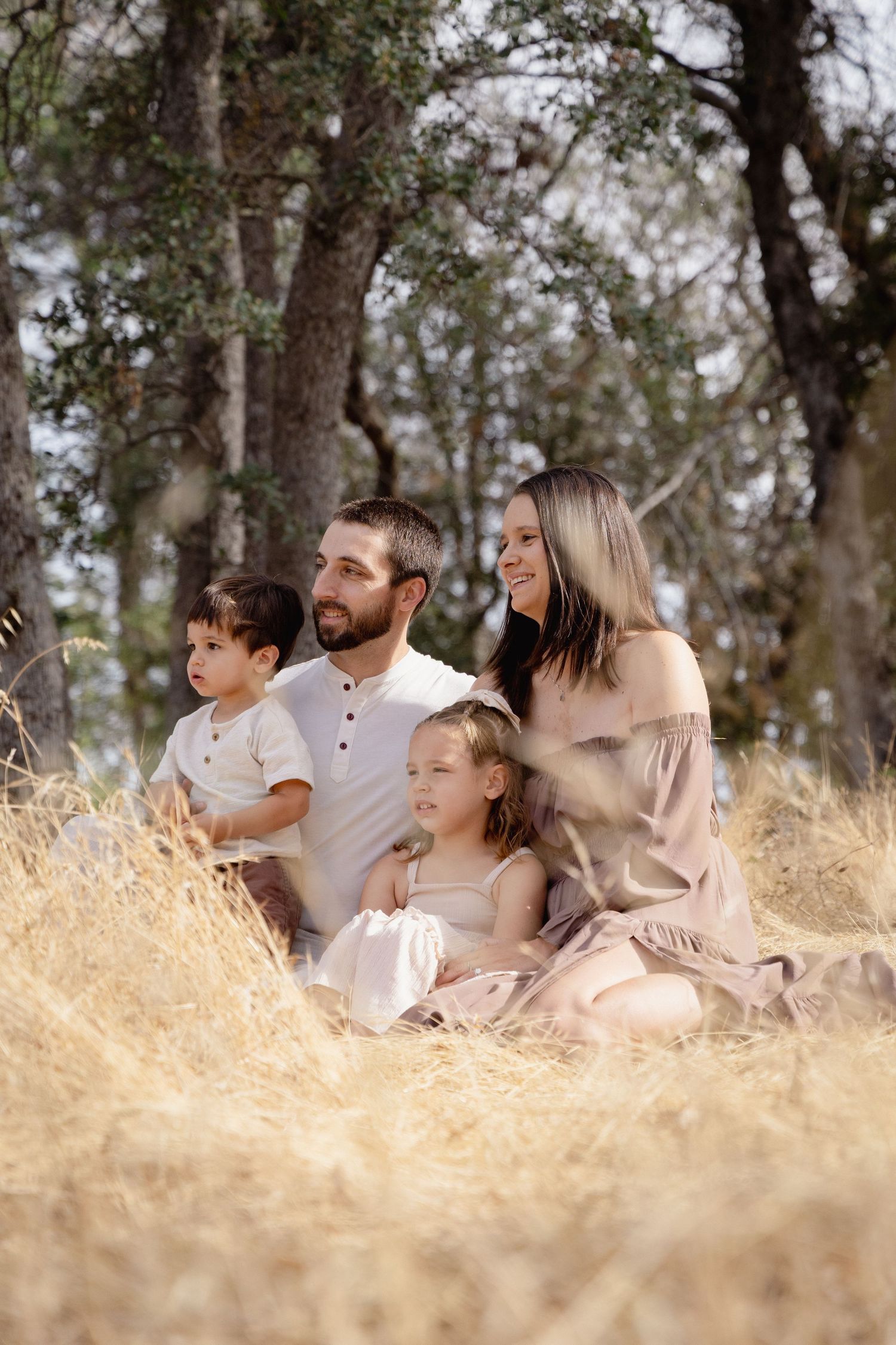 Family of four sitting in golden grass among trees, parents smiling with two young children on a sunny day.