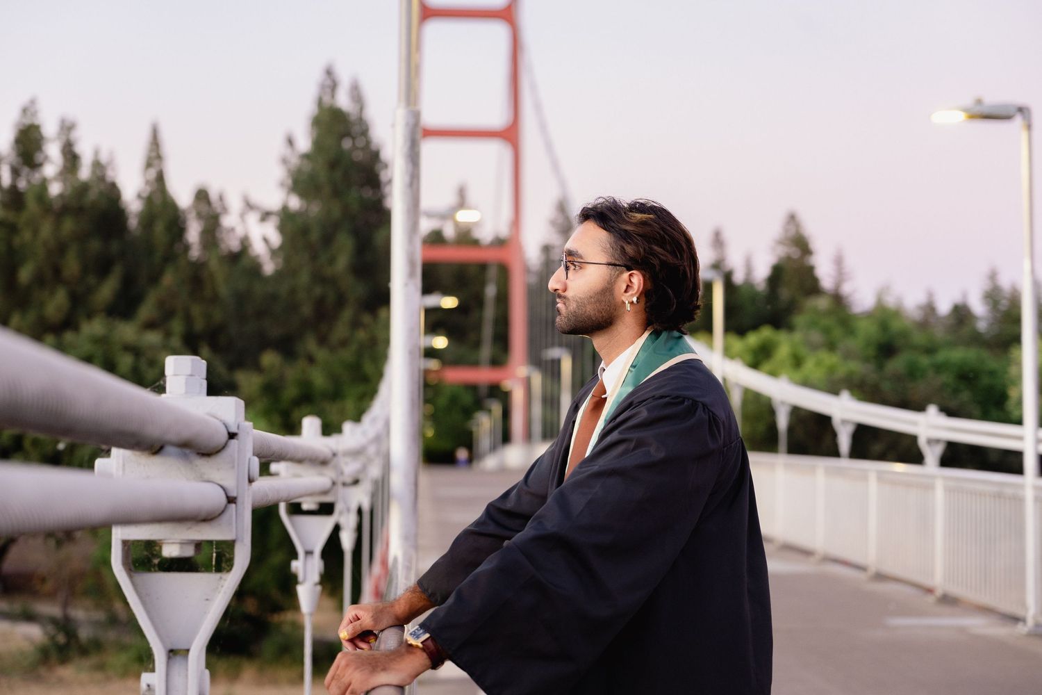Graduate in cap and gown stands on a bridge, gazing thoughtfully into the distance with trees in the background.