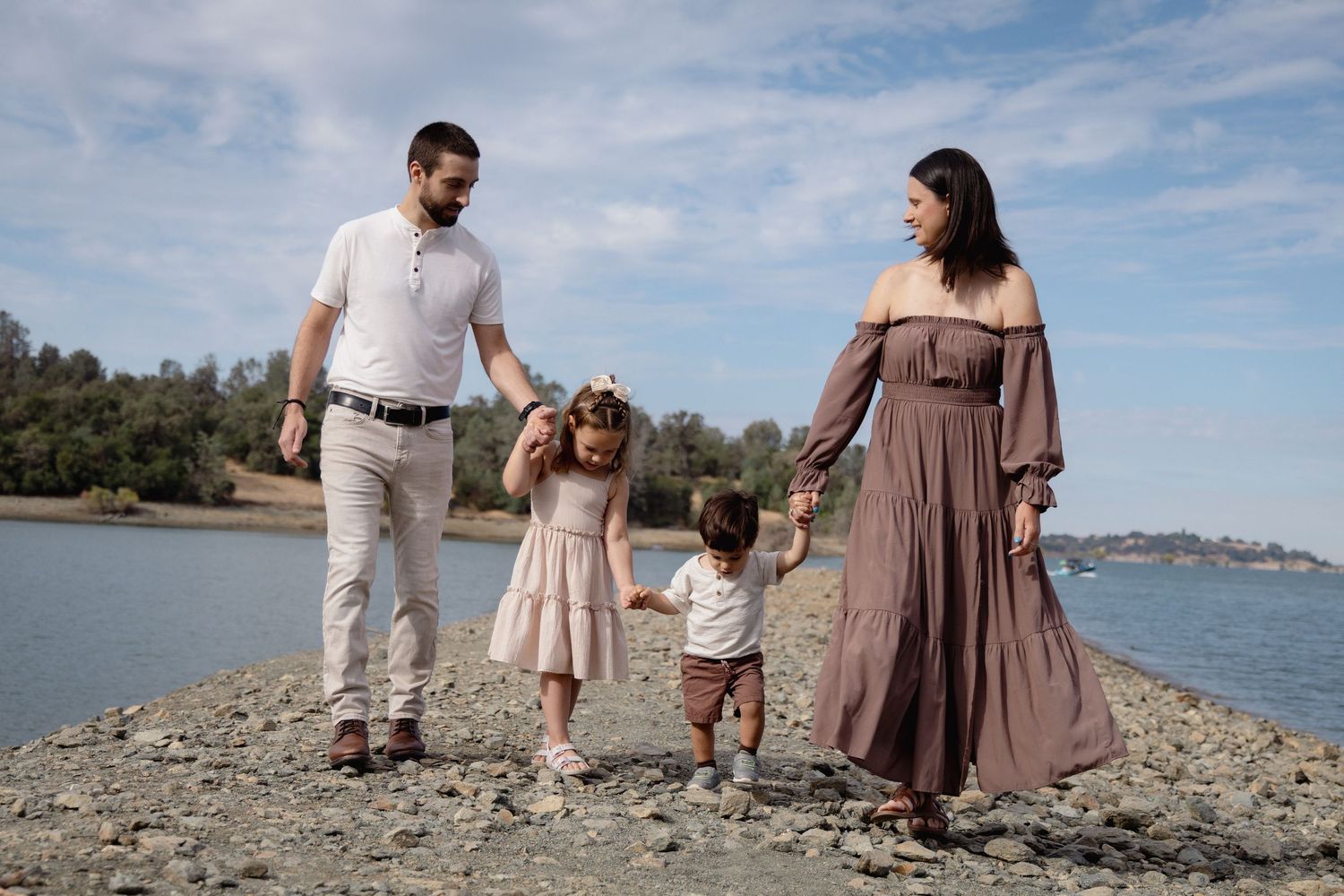 Family of four walking on rocky shoreline, parents holding hands with young children near calm blue water.