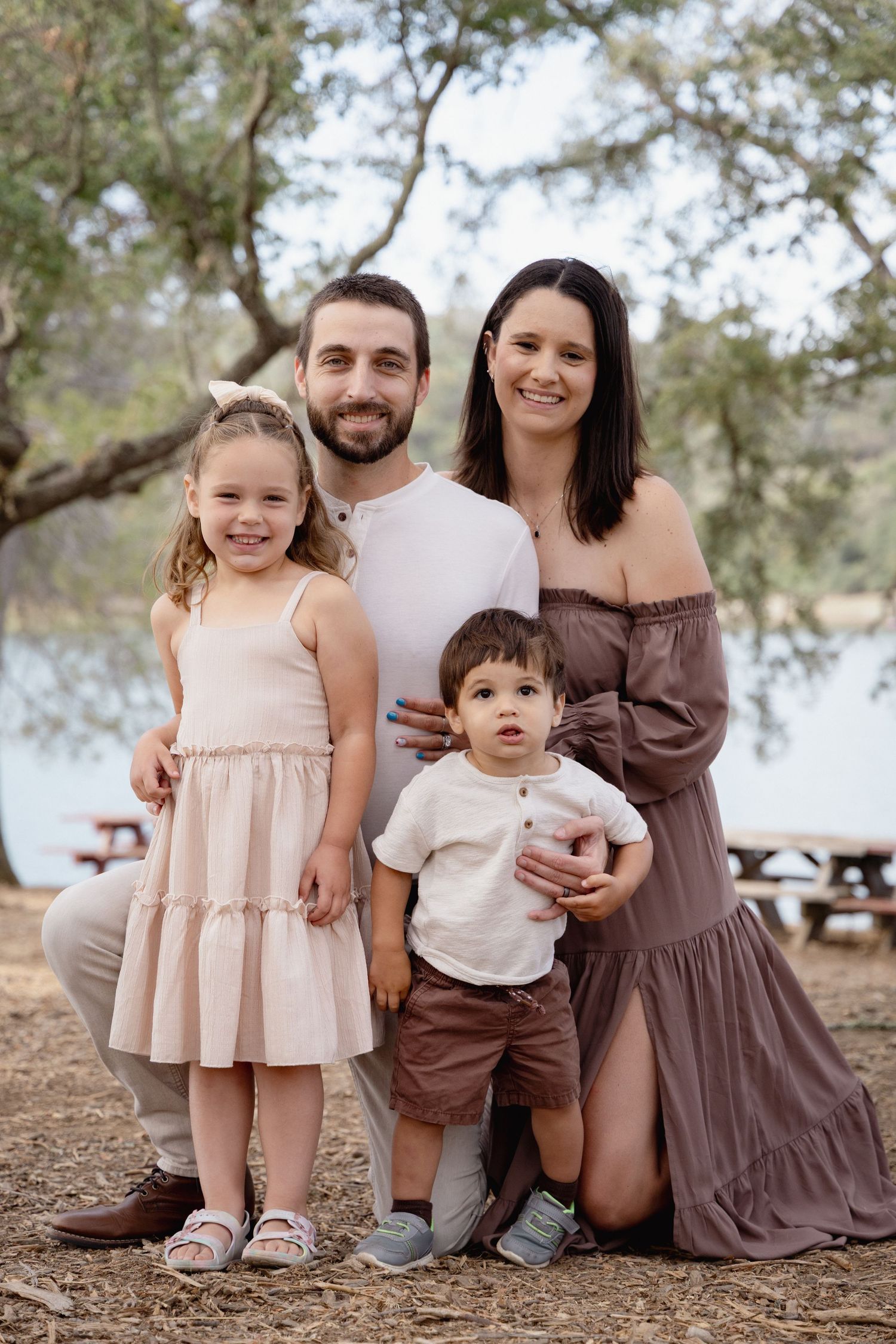 Happy family of four posing outdoors near a lake, parents with young daughter in pink dress and toddler son.