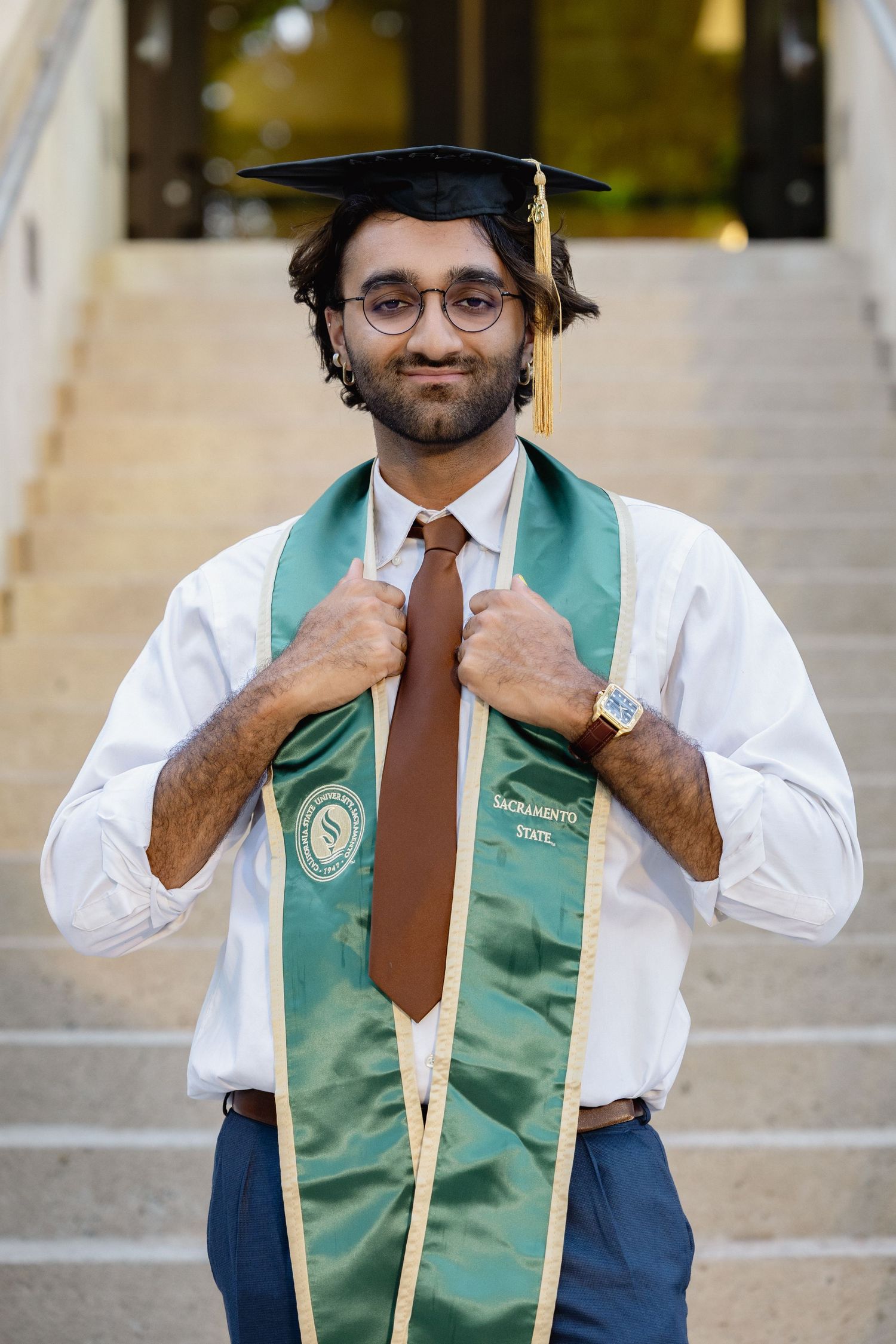 Smiling graduate wearing mortarboard and green Sacramento State stole stands on outdoor steps.