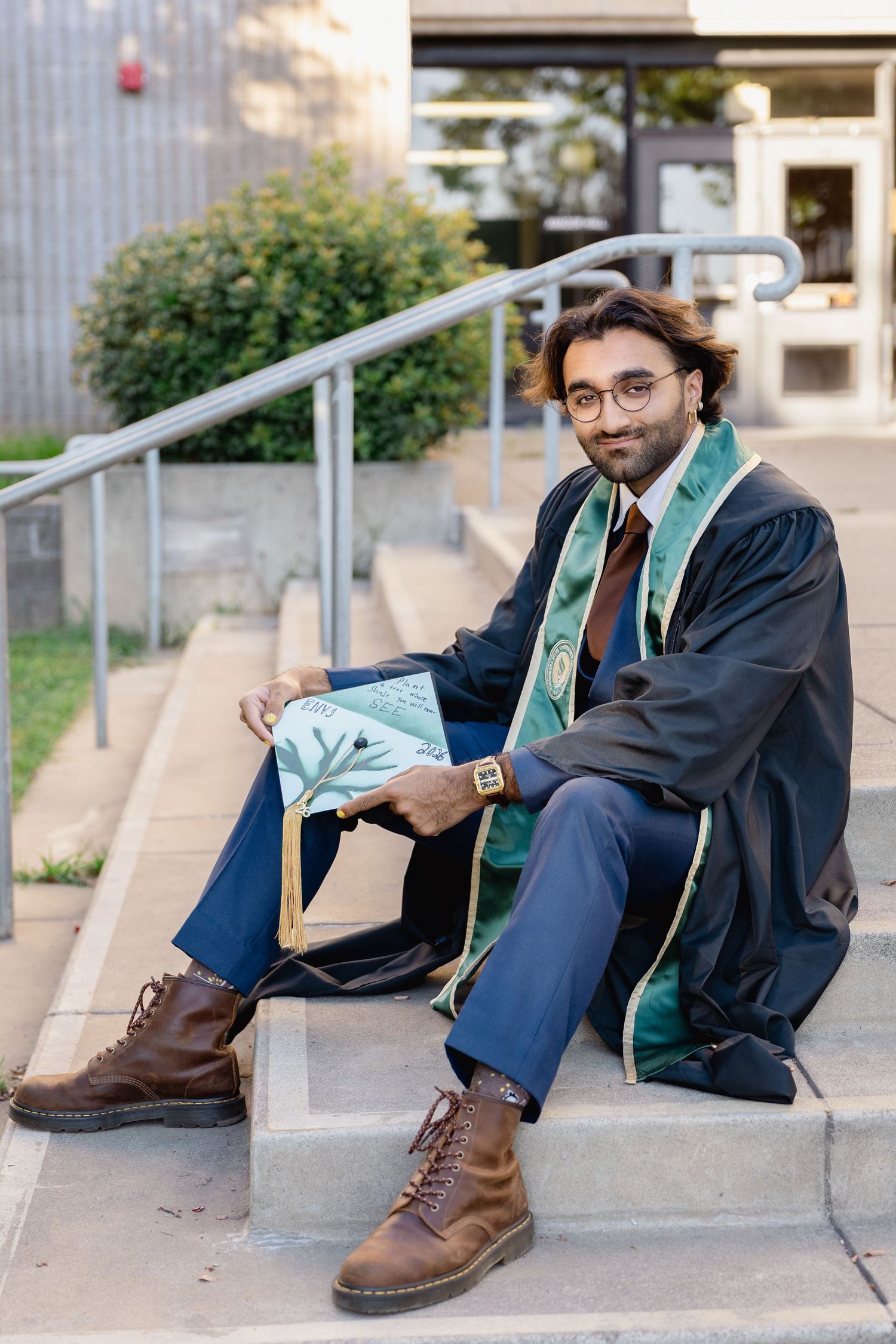 Graduate in black robe and green Sacramento State stole sits on steps holding mortarboard cap.