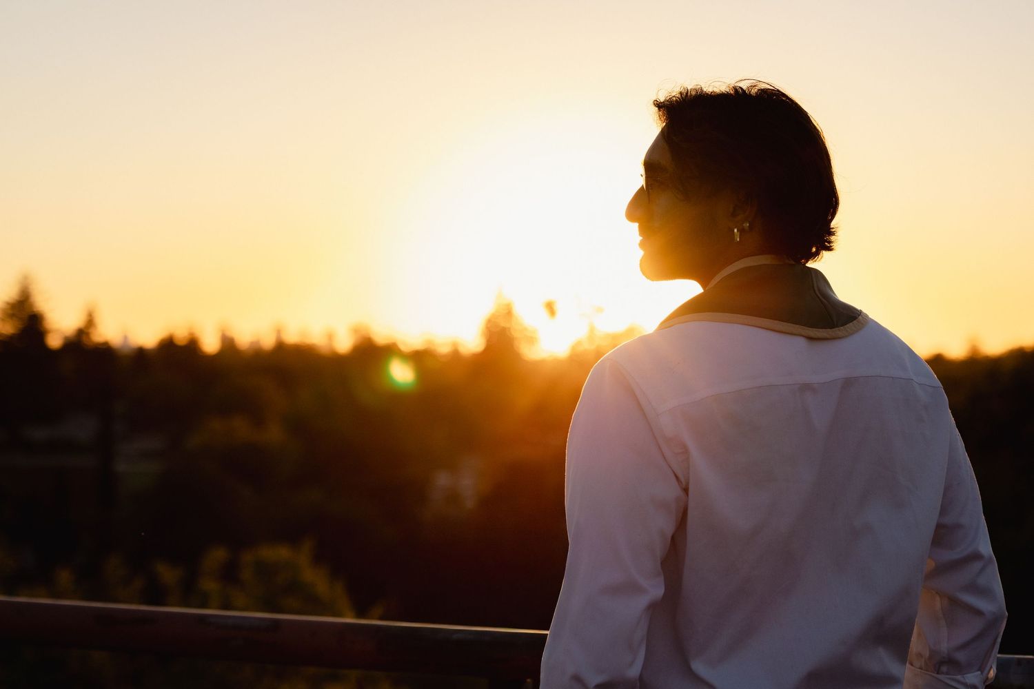 Person in white shirt gazing at golden sunset over city skyline from elevated viewpoint.