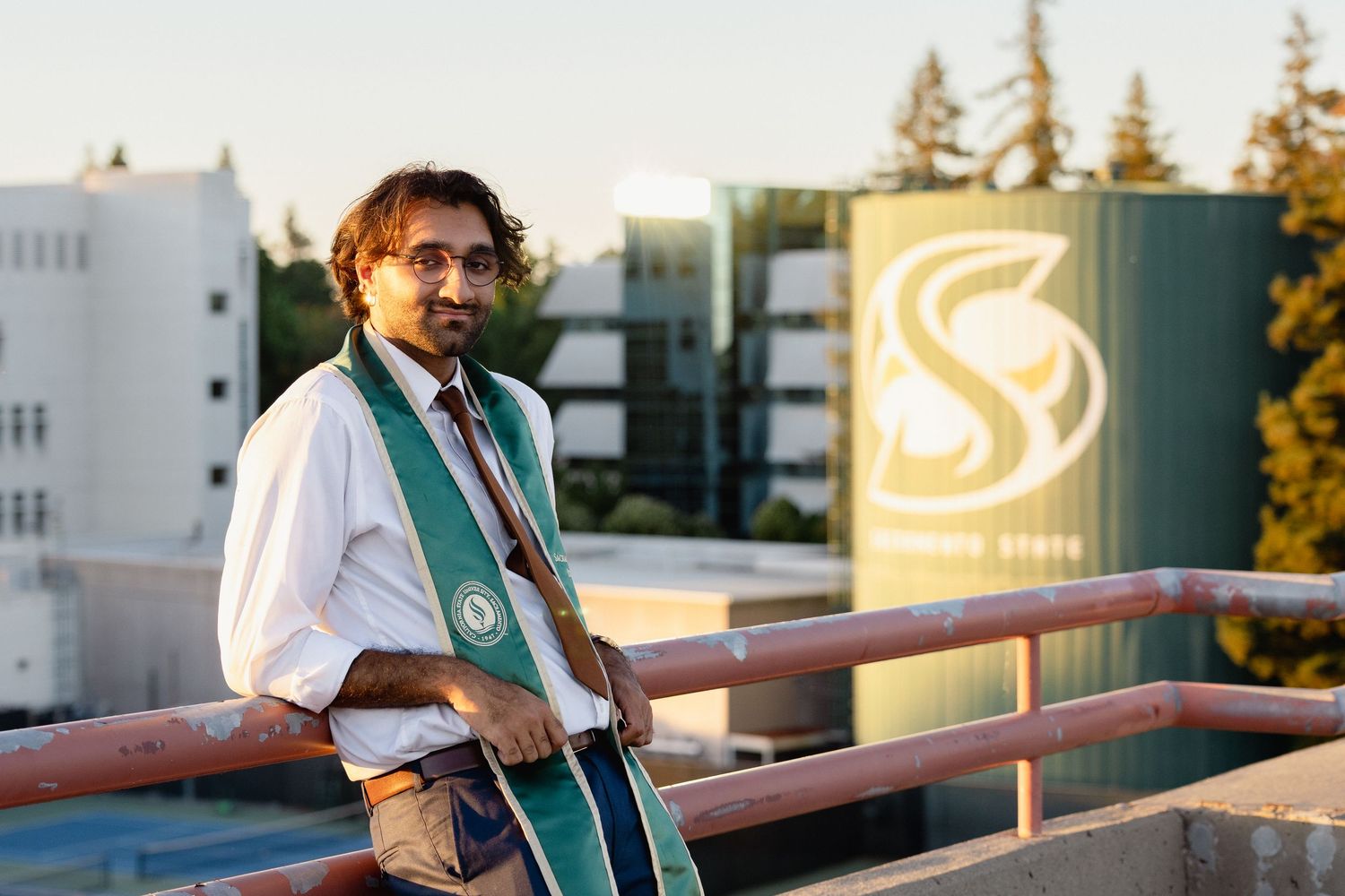 Graduate student in cap and gown sash poses on rooftop with Sonoma State University sign in background.