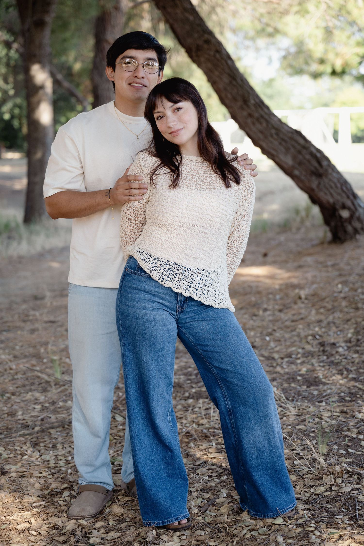 A couple poses together outdoors among trees, wearing casual cream tops and blue jeans, smiling warmly.
