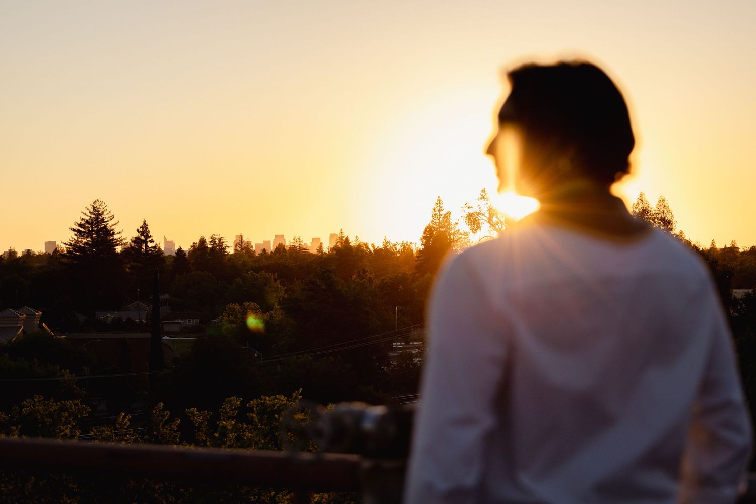 Person in white hoodie watching a golden sunset over tree-lined horizon in serene evening light.