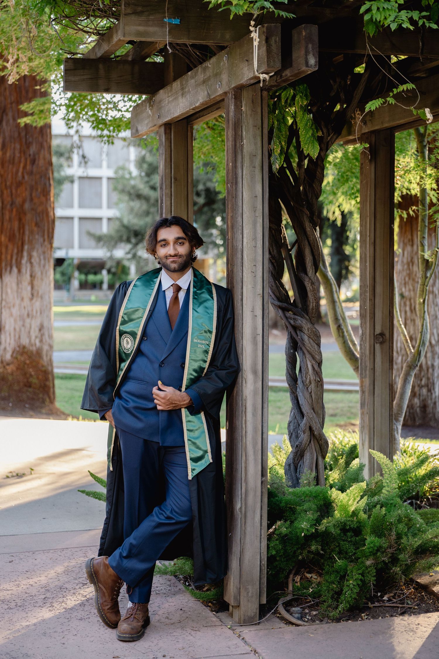 Young man in graduation gown and stole poses confidently under wooden pergola in blue suit outdoors.