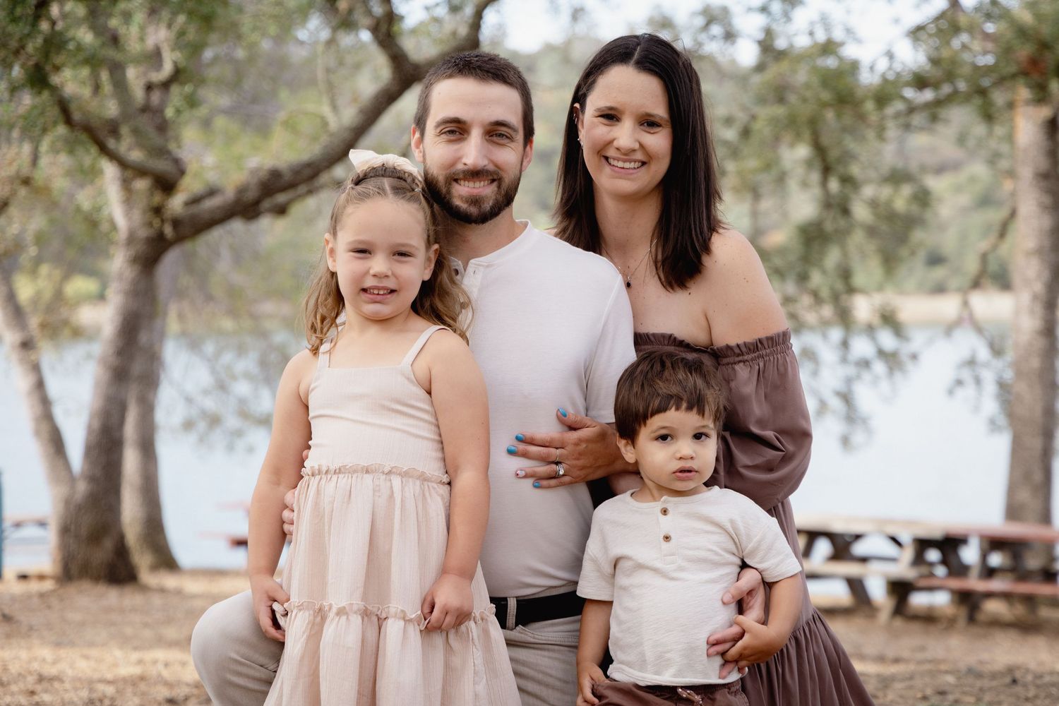 Happy family of four posing outdoors among trees, parents with two young children smiling in casual attire.