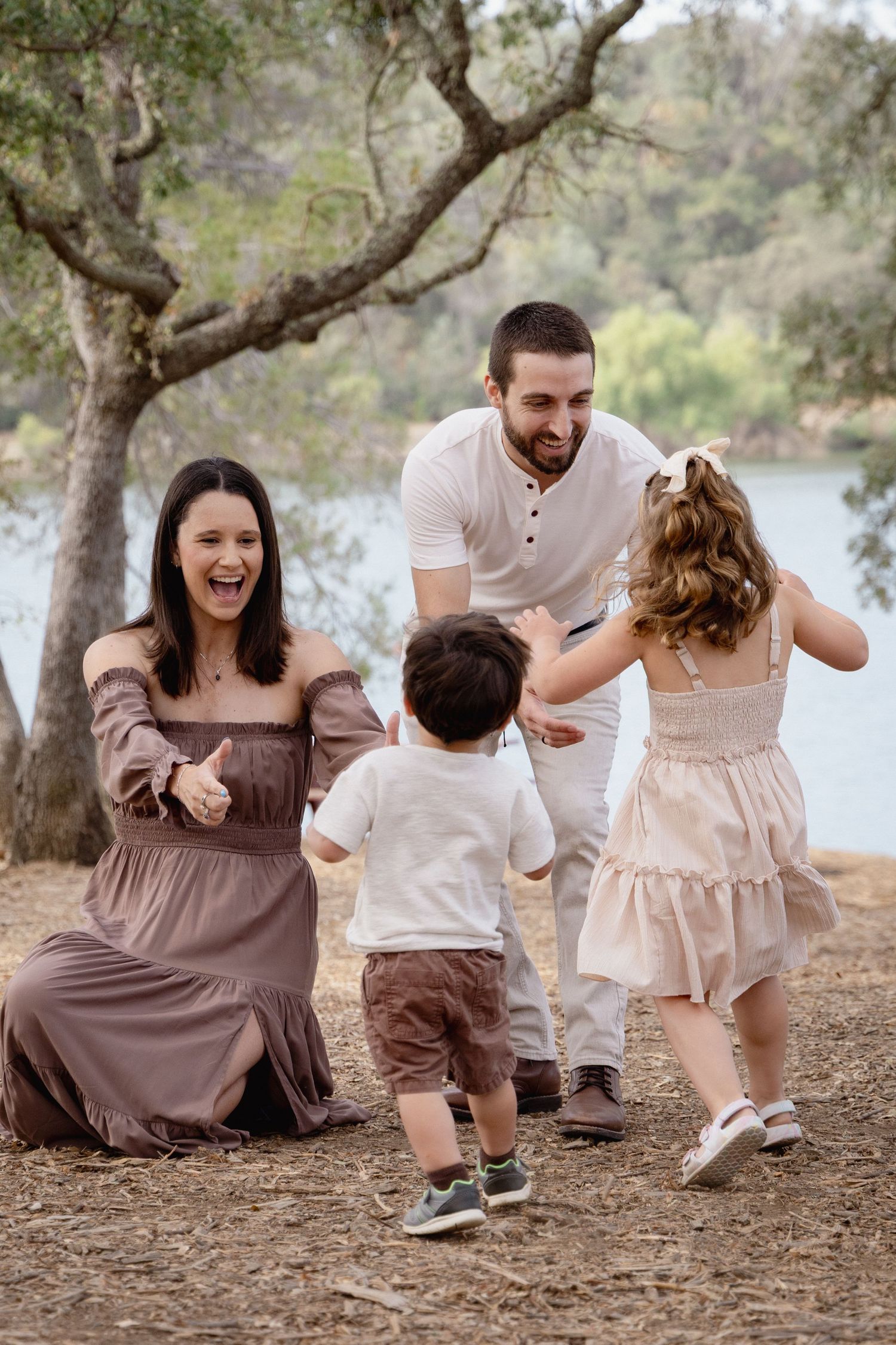 Happy family of four playing outdoors near a lake, parents laughing as young children run toward them.