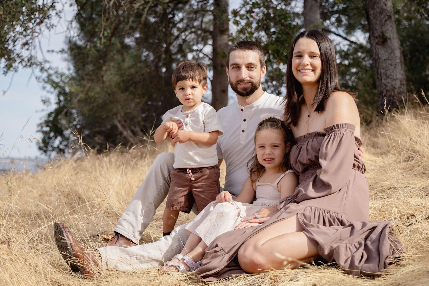 Family of four sitting in golden grass field, parents with toddler son and young daughter, outdoor portrait.