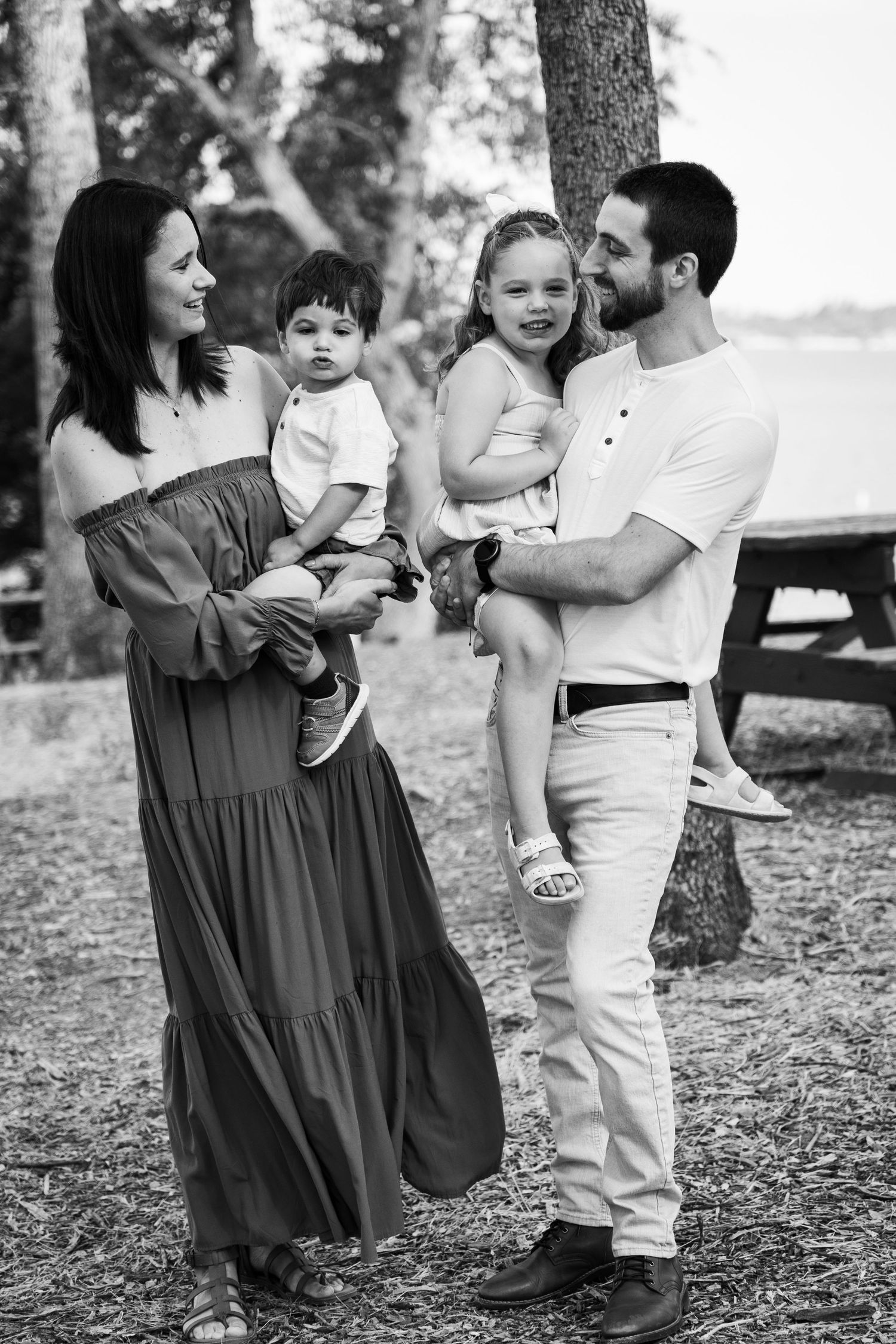 Black and white family portrait of parents holding two young children outdoors near trees and a lake.