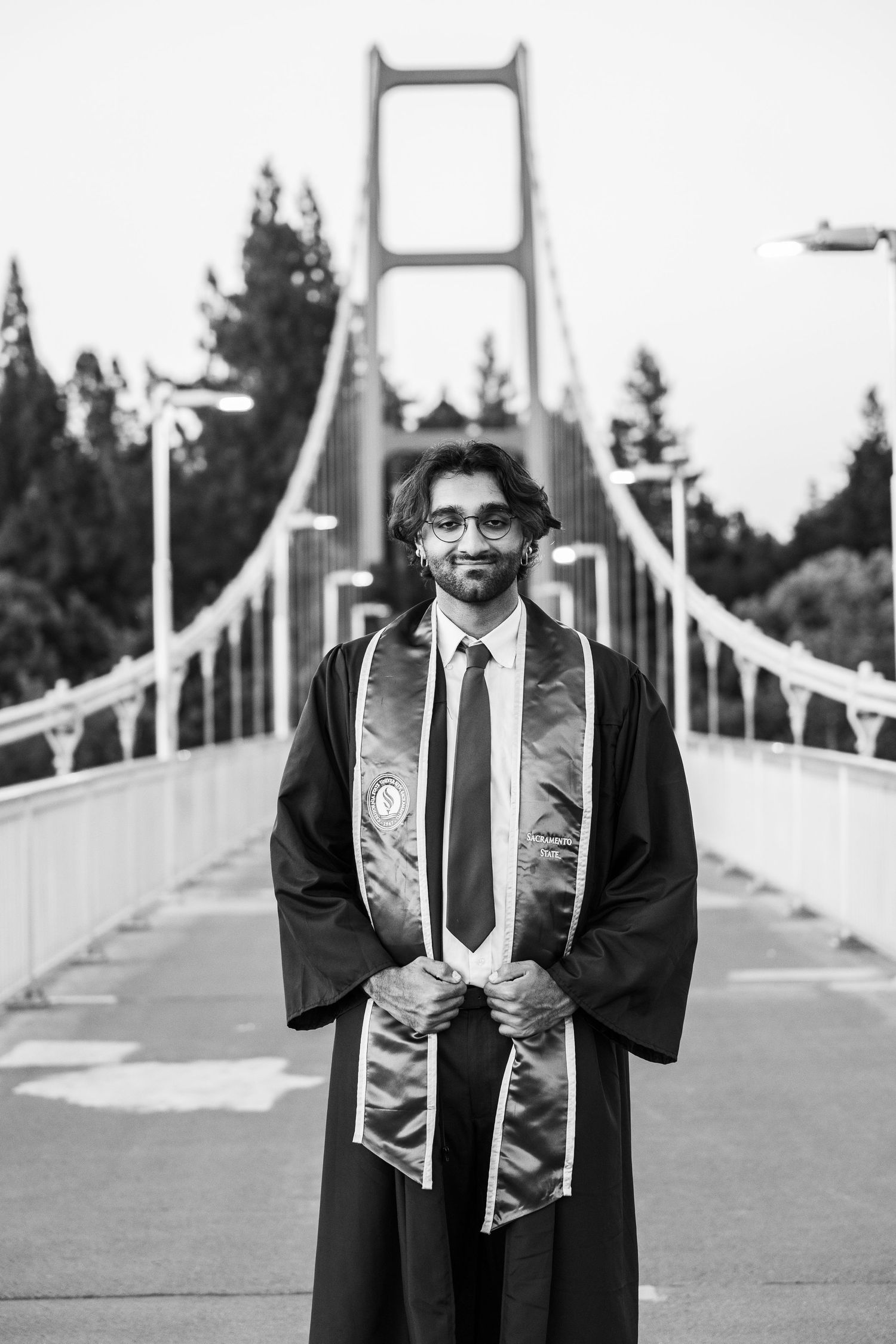 Black and white photo of graduate in robe and stole standing on suspension bridge.