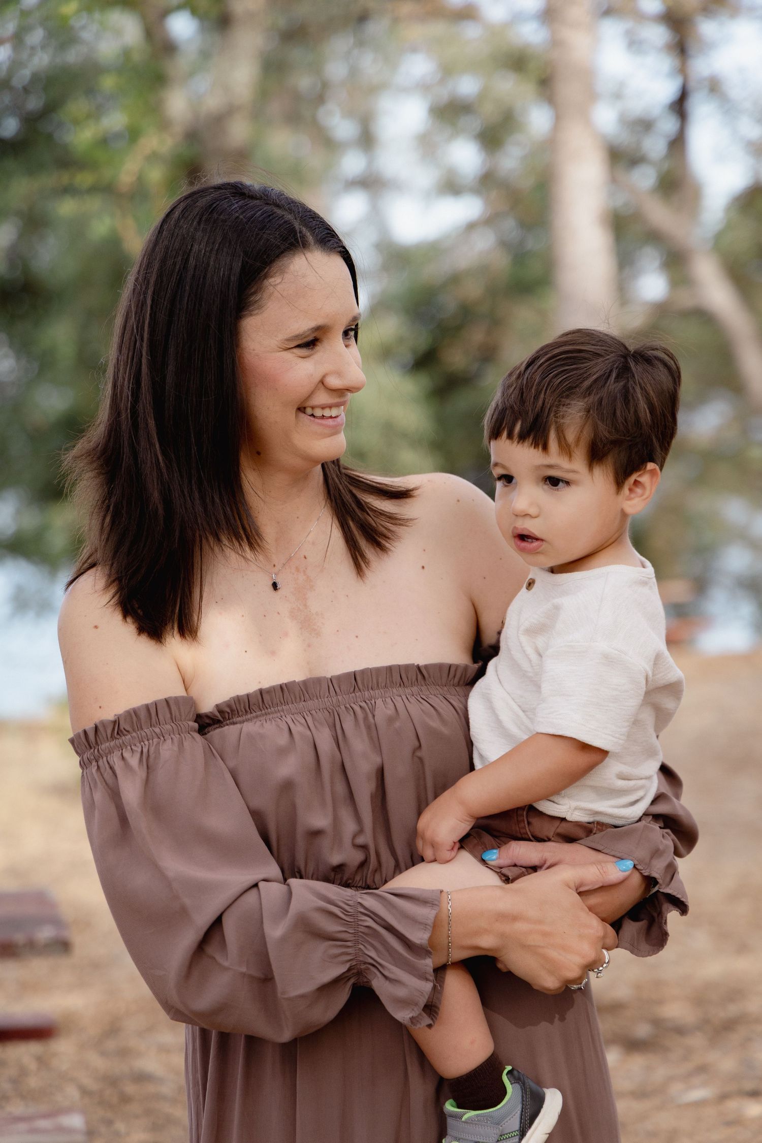 A smiling woman in an off-shoulder brown dress holds a young toddler boy outdoors near trees.