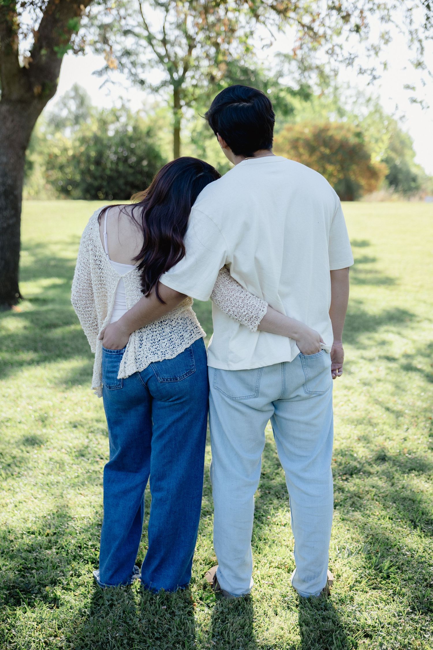 Couple embracing from behind in a sunny park, wearing casual white and blue outfits, standing on green grass.