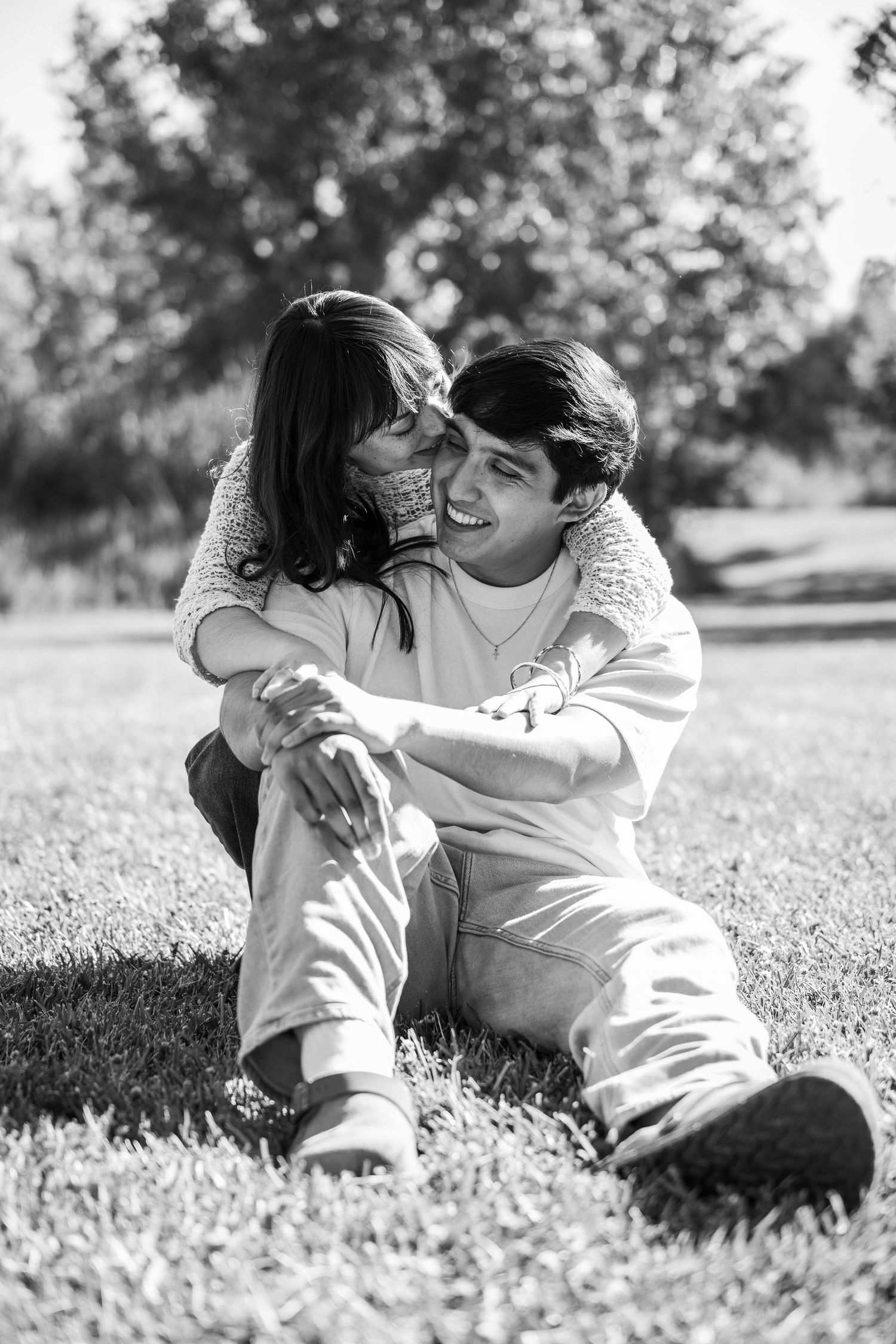Black and white photo of a happy couple sitting on grass, woman kissing man on cheek in a sunny park.