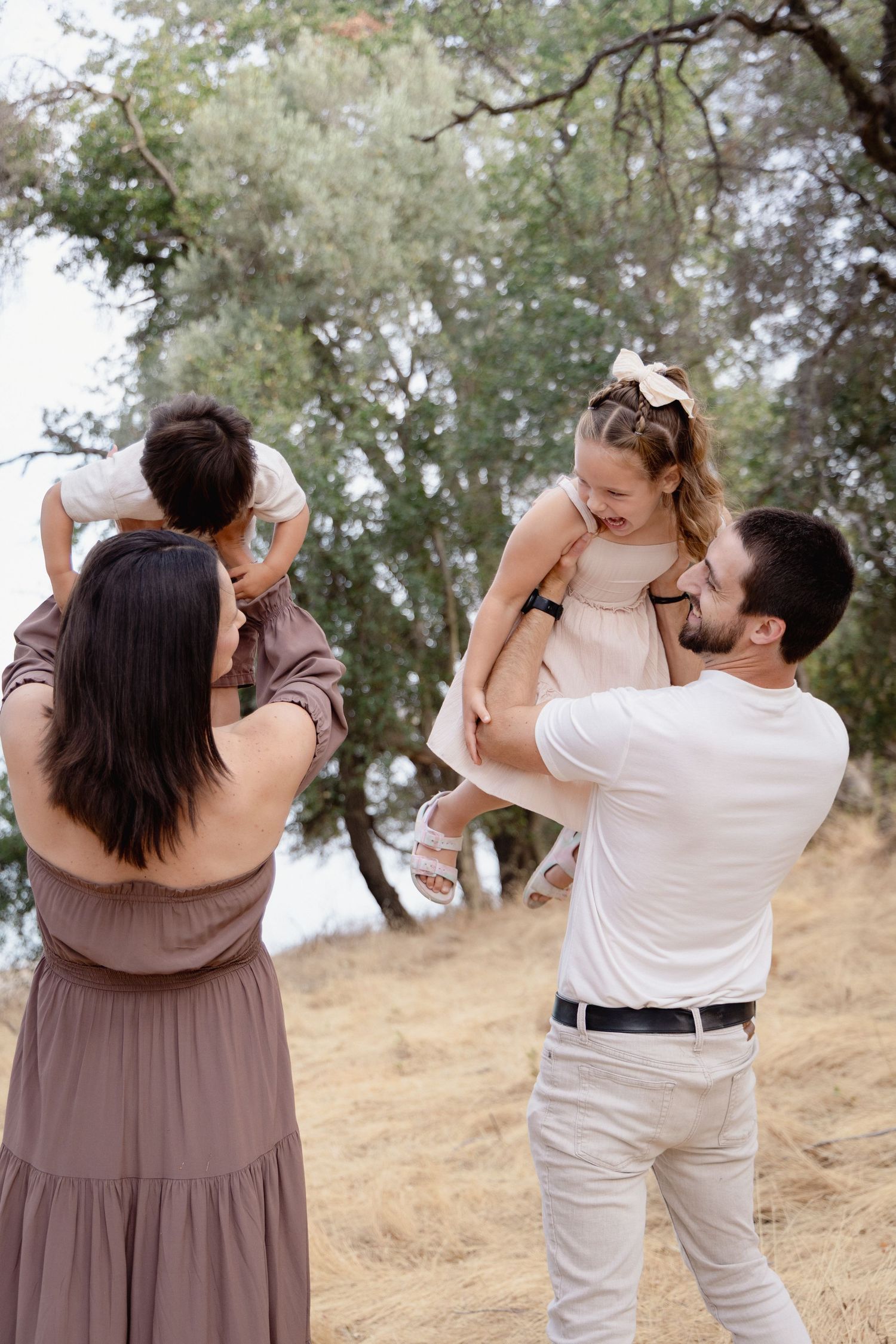 A happy family of four plays outdoors, with parents lifting two young children among trees on a sunny day.