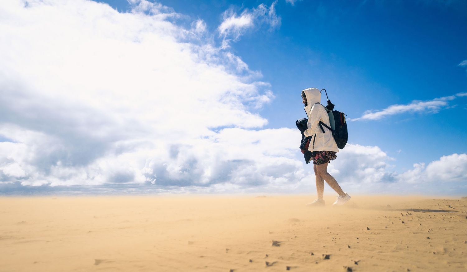 Person with backpack walking across vast sandy desert under dramatic cloudy blue sky.