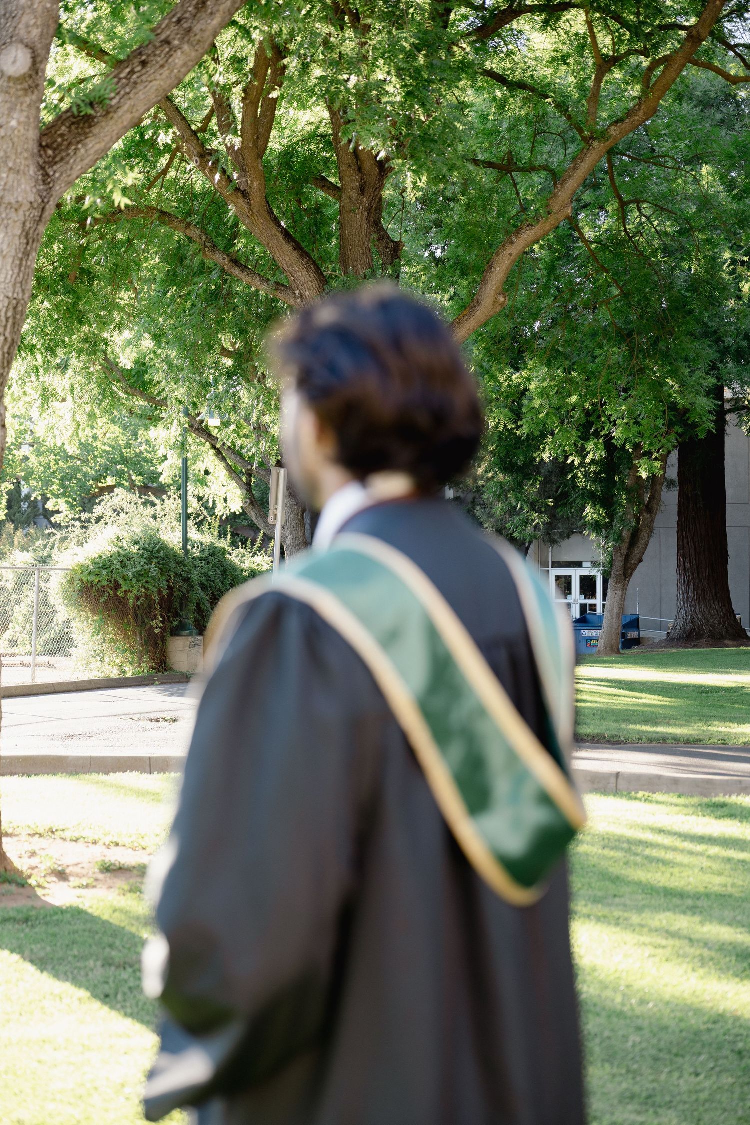 Graduate in cap and gown standing outdoors on a green campus lawn with trees in the background.
