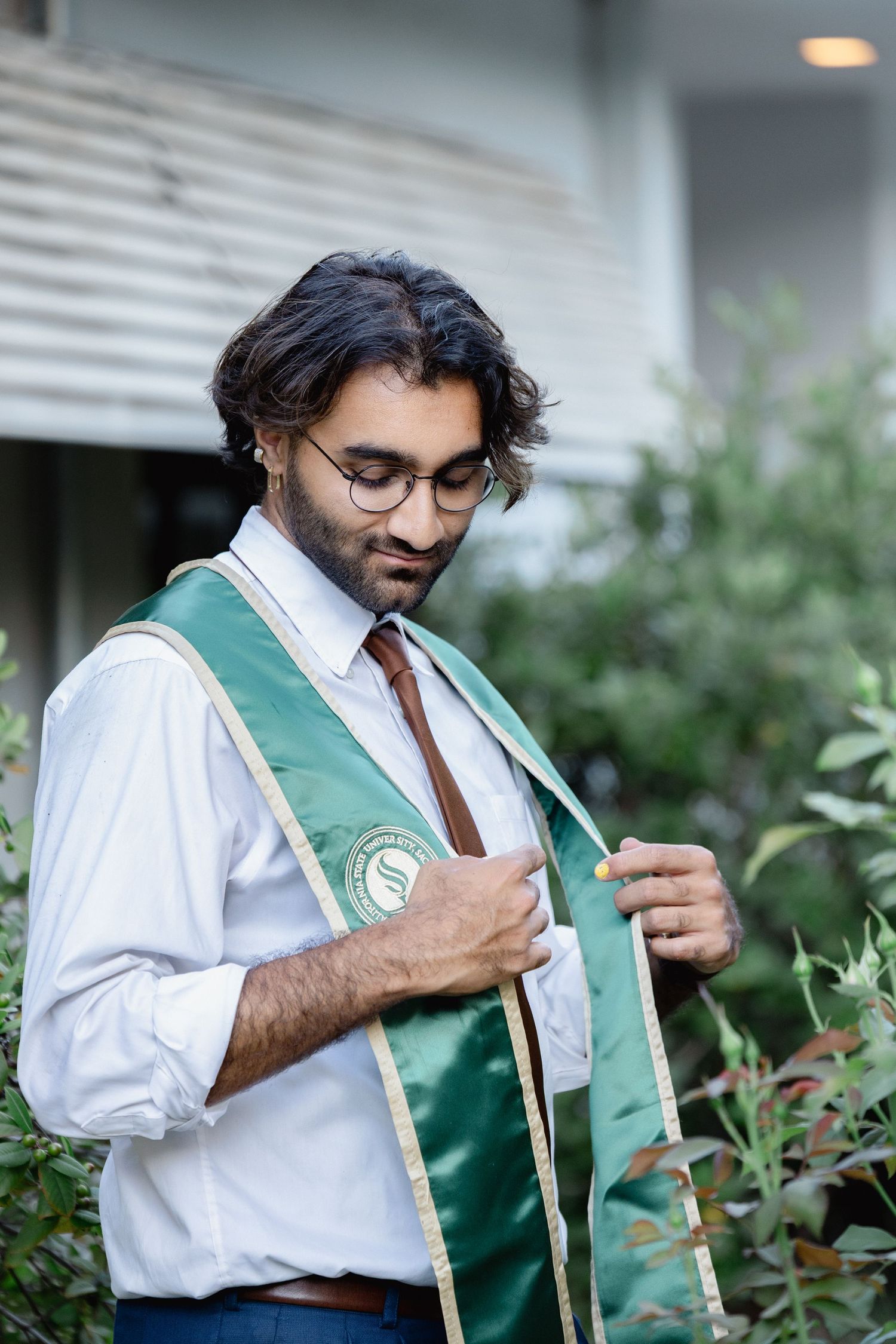 Young man in glasses and green graduation sash adjusting his attire outdoors near green bushes.