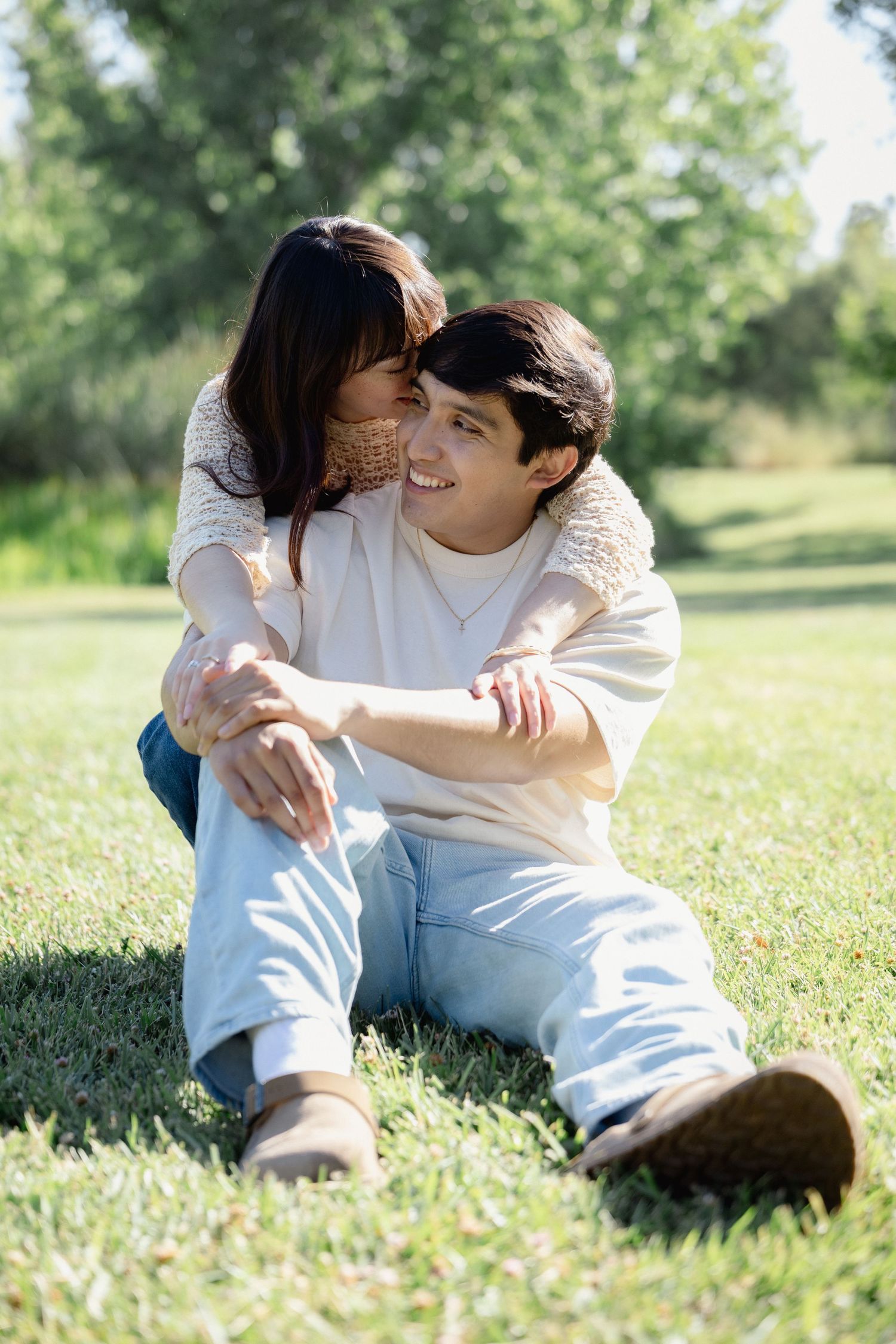 Young couple sitting on grass in park, woman hugging man from behind, smiling on sunny day.