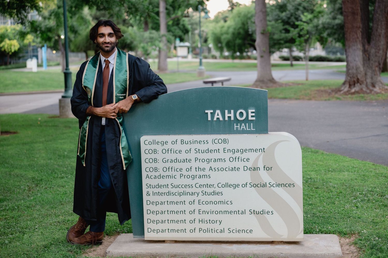 Graduate in cap and gown poses proudly beside Tahoe Hall sign at college campus on graduation day.