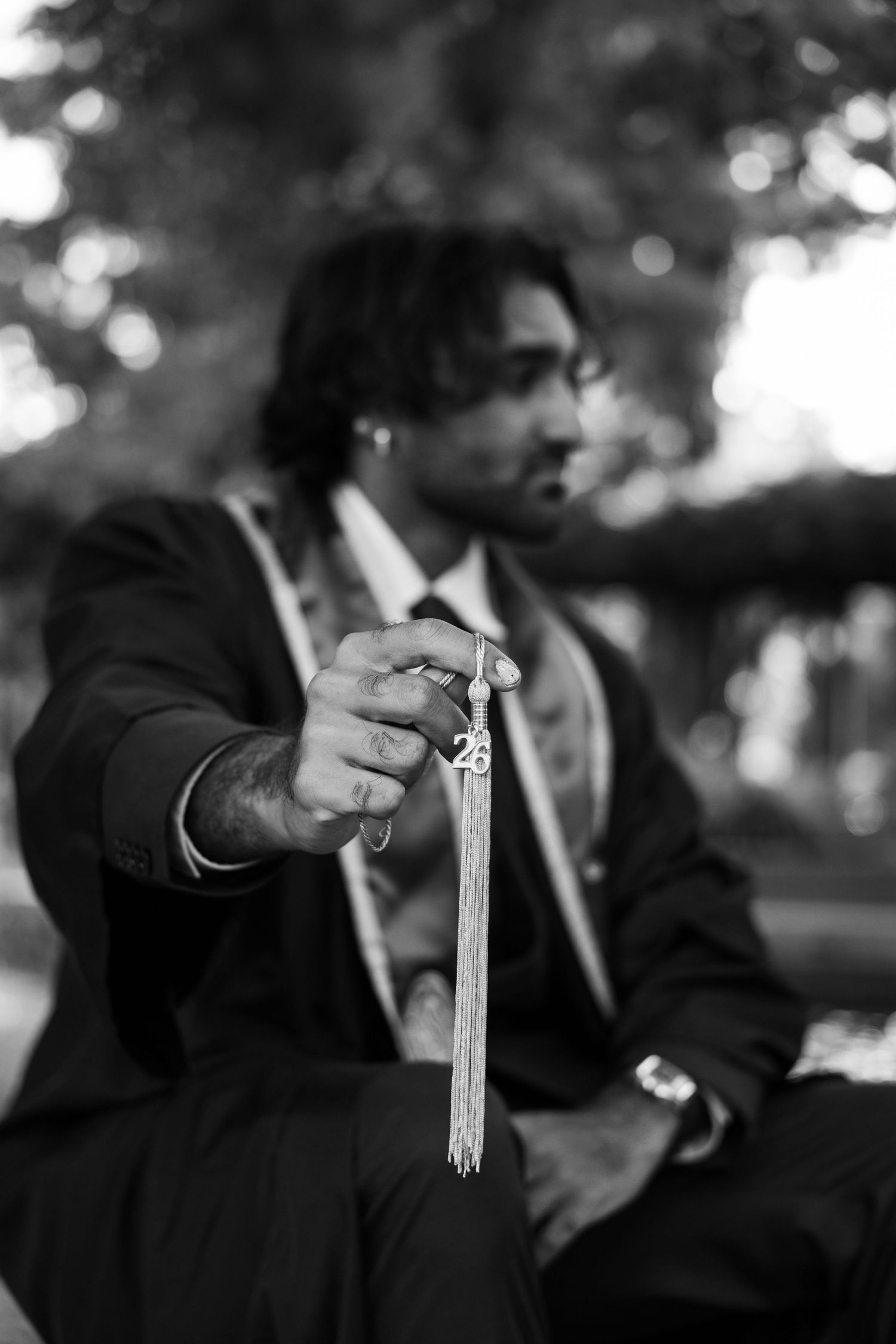 Black and white photo of a man in a suit holding graduation tassel toward camera outdoors.