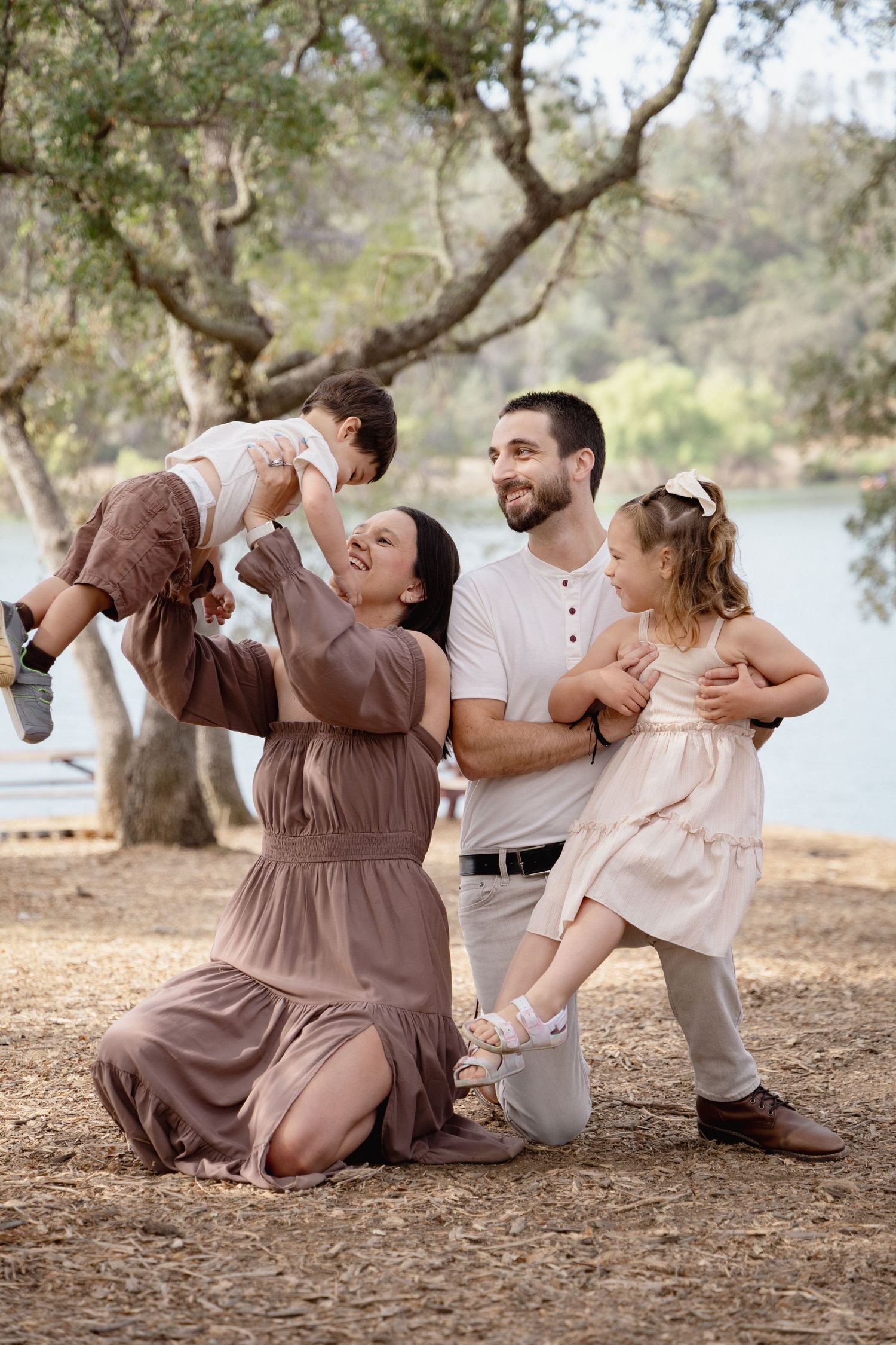 Happy family playing outdoors near a lake, parents lifting children among trees in a warm, natural setting.