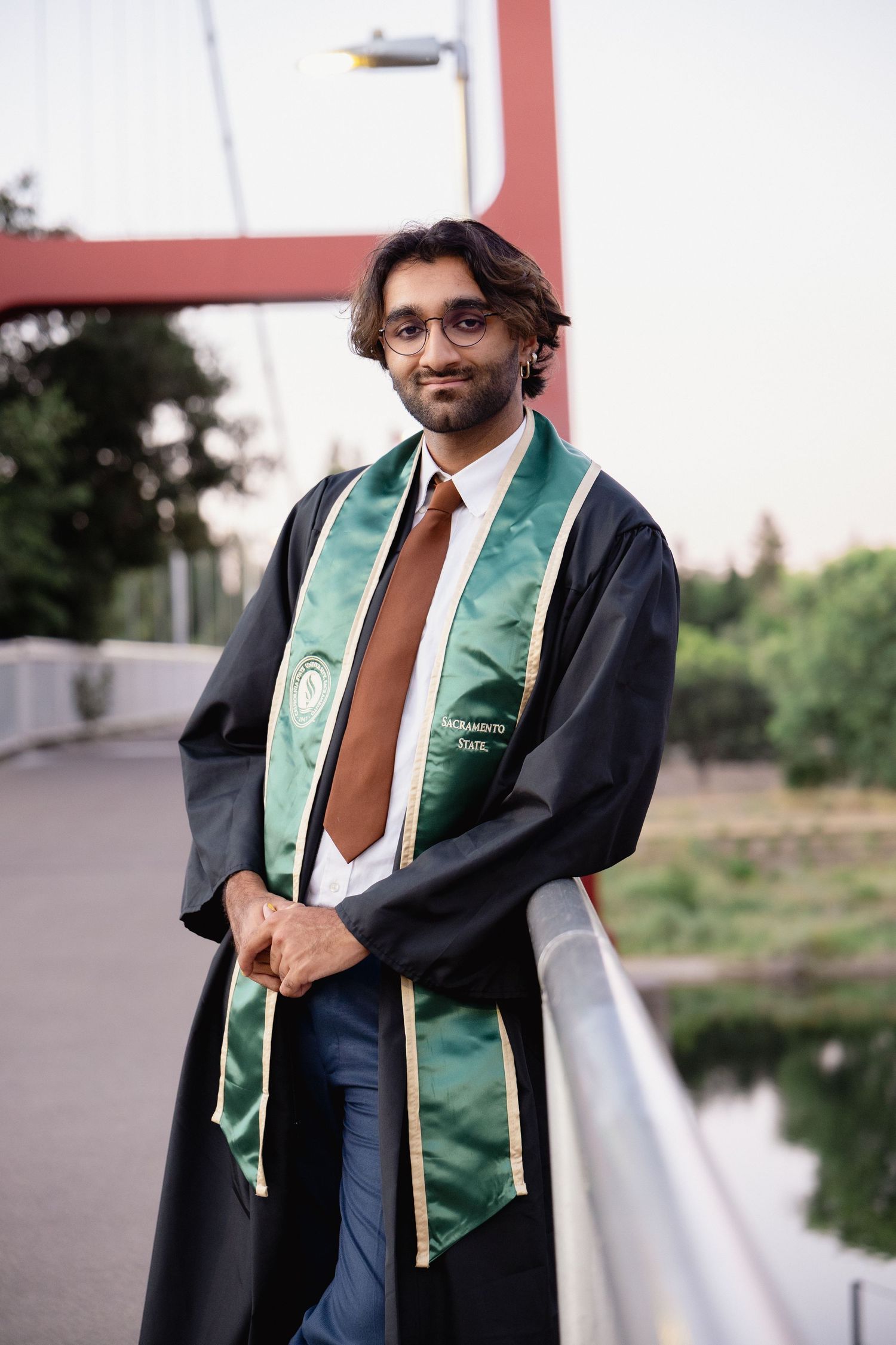 Graduate in black robe and green Sacramento State stole poses on bridge at dusk.