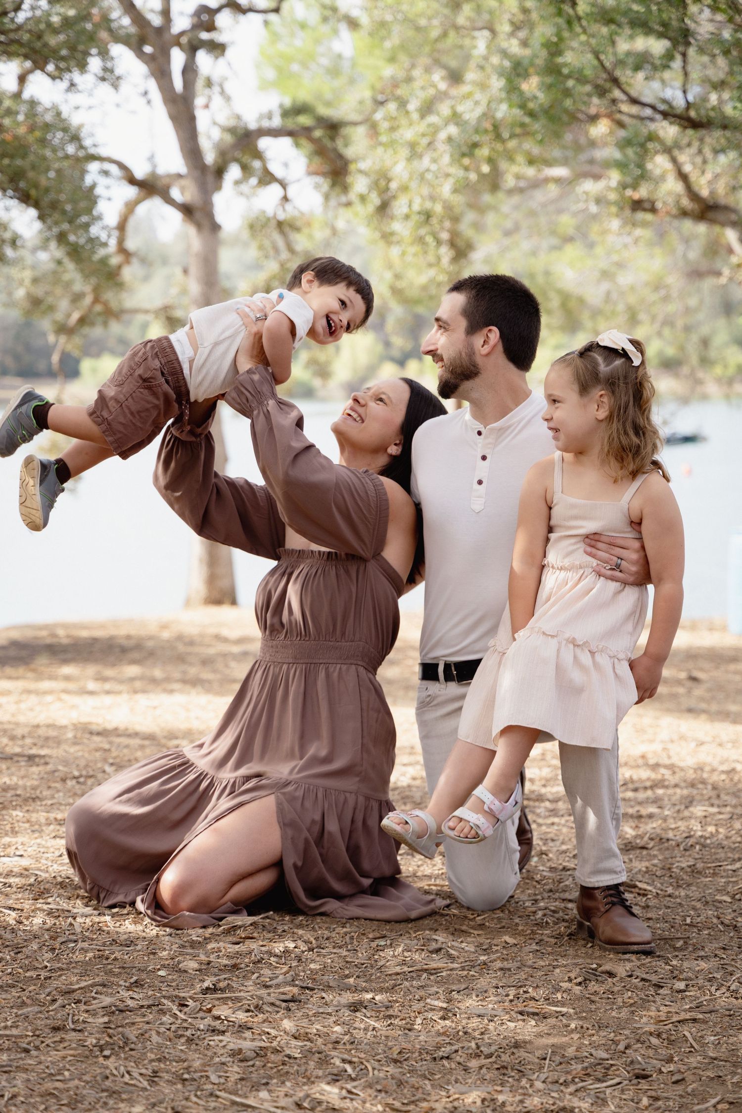 Happy family of four enjoying a sunny day at the park, with mom lifting toddler son while dad and daughter smile nearby.