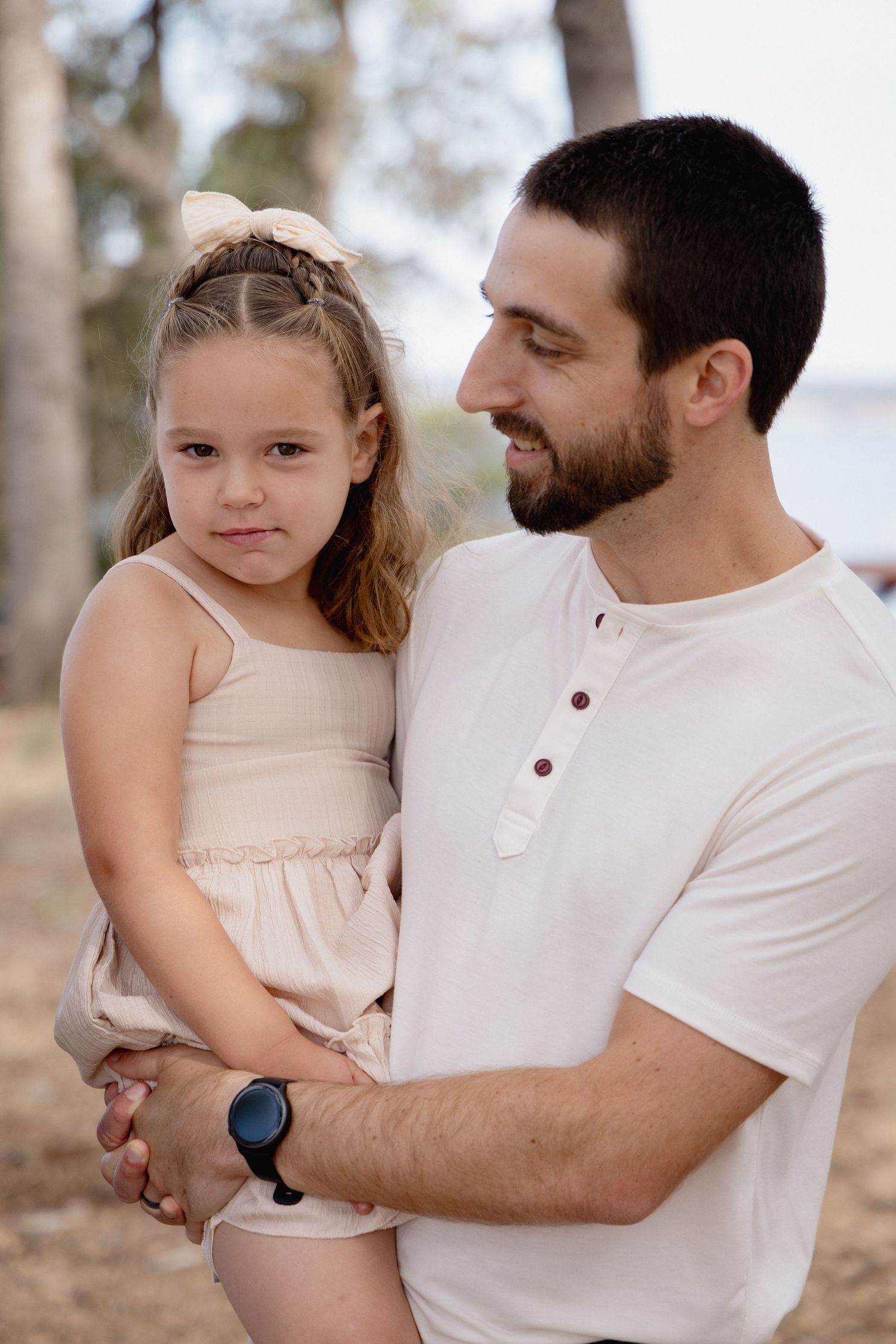 A bearded man in a white shirt holds a young girl in a beige dress outdoors among trees.