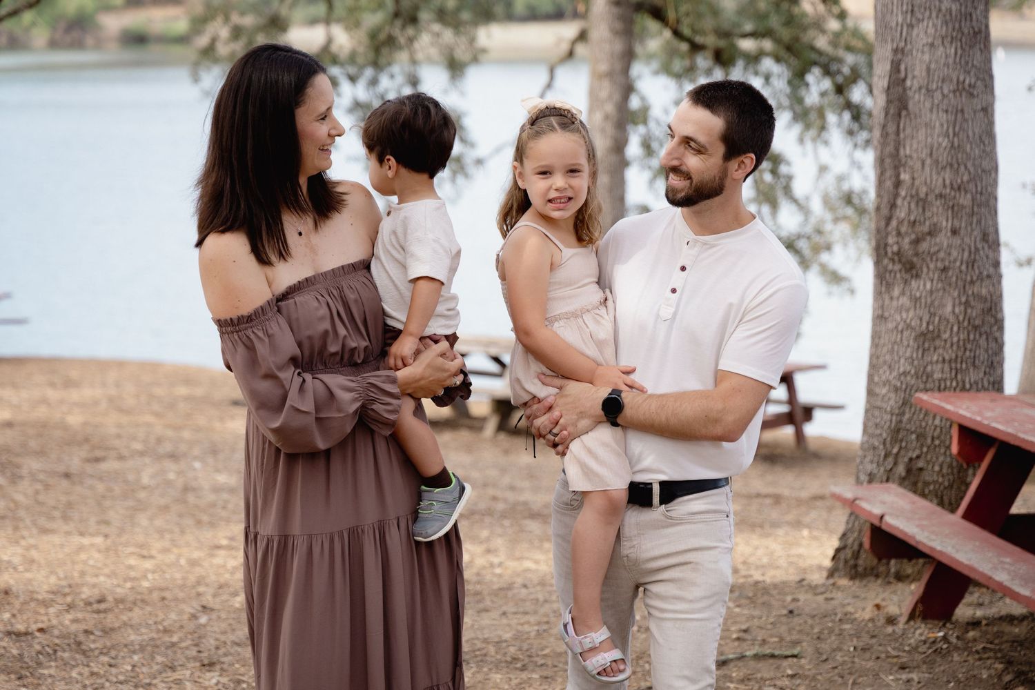 Family of four enjoying a sunny day by the lake, with parents holding two young children near trees.