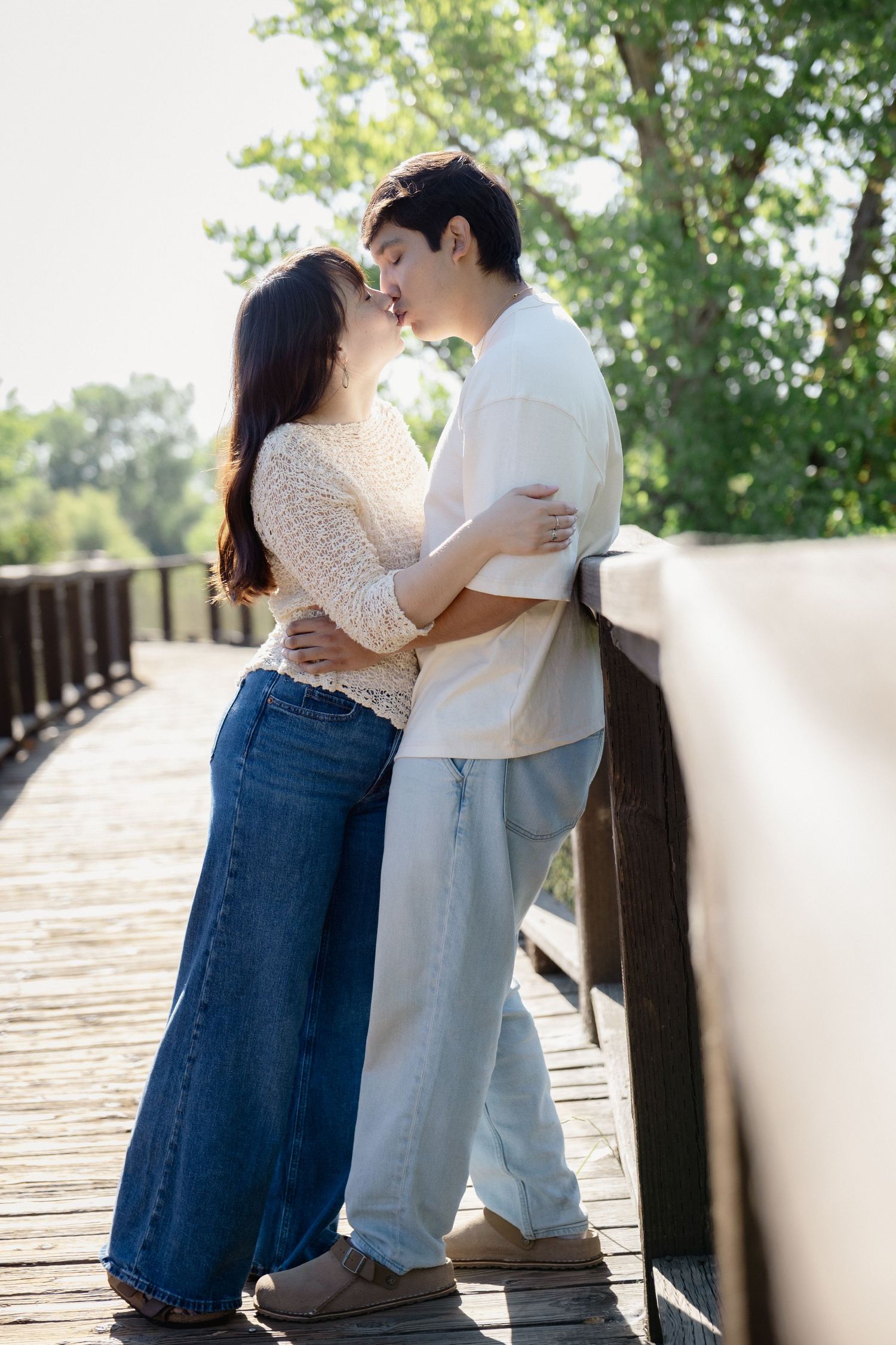 Couple sharing a romantic kiss on a wooden bridge surrounded by lush green trees on a sunny day.