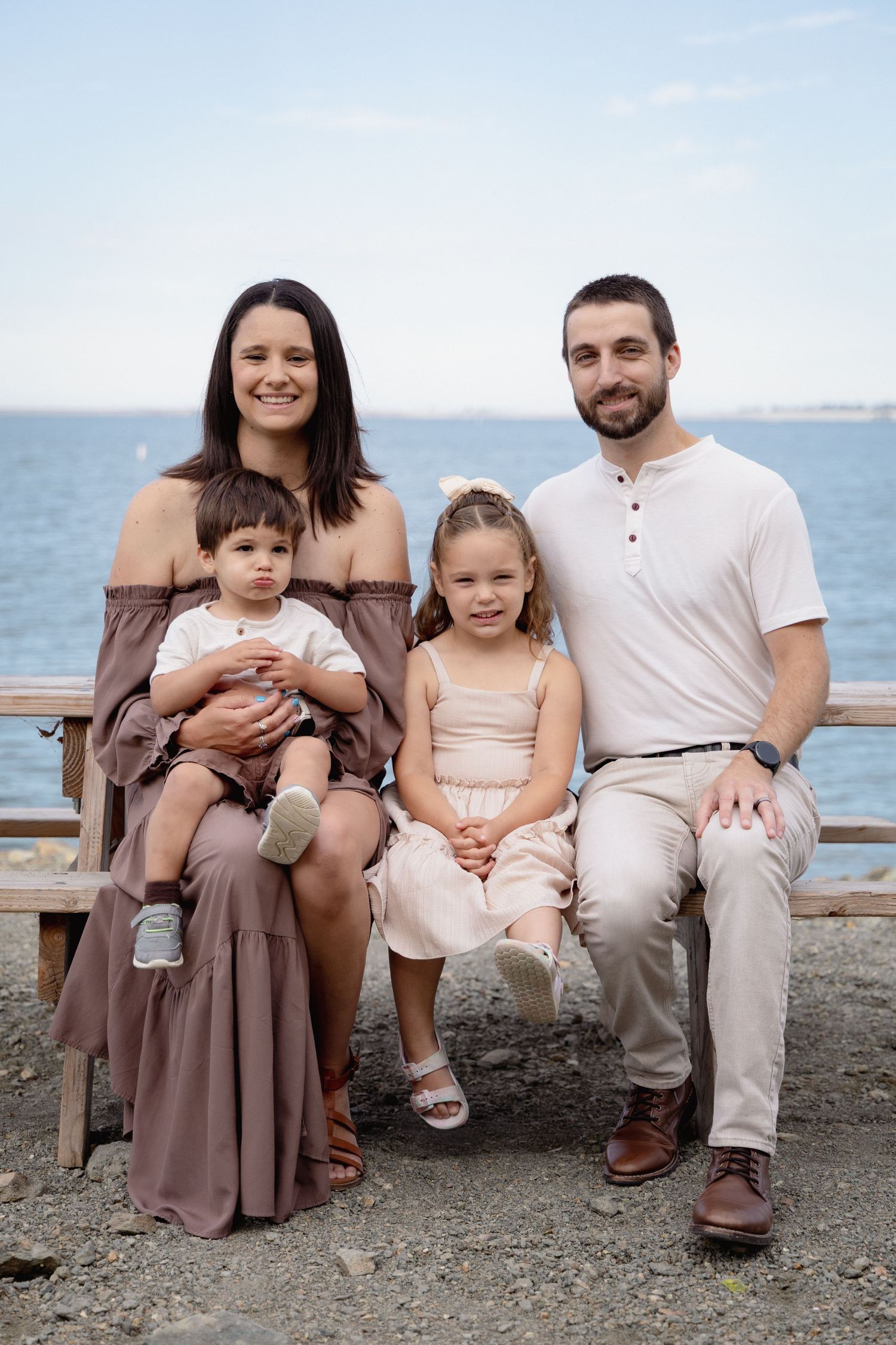 Happy family of four posing on a bench by the ocean, parents with toddler and young girl in coordinated outfits.