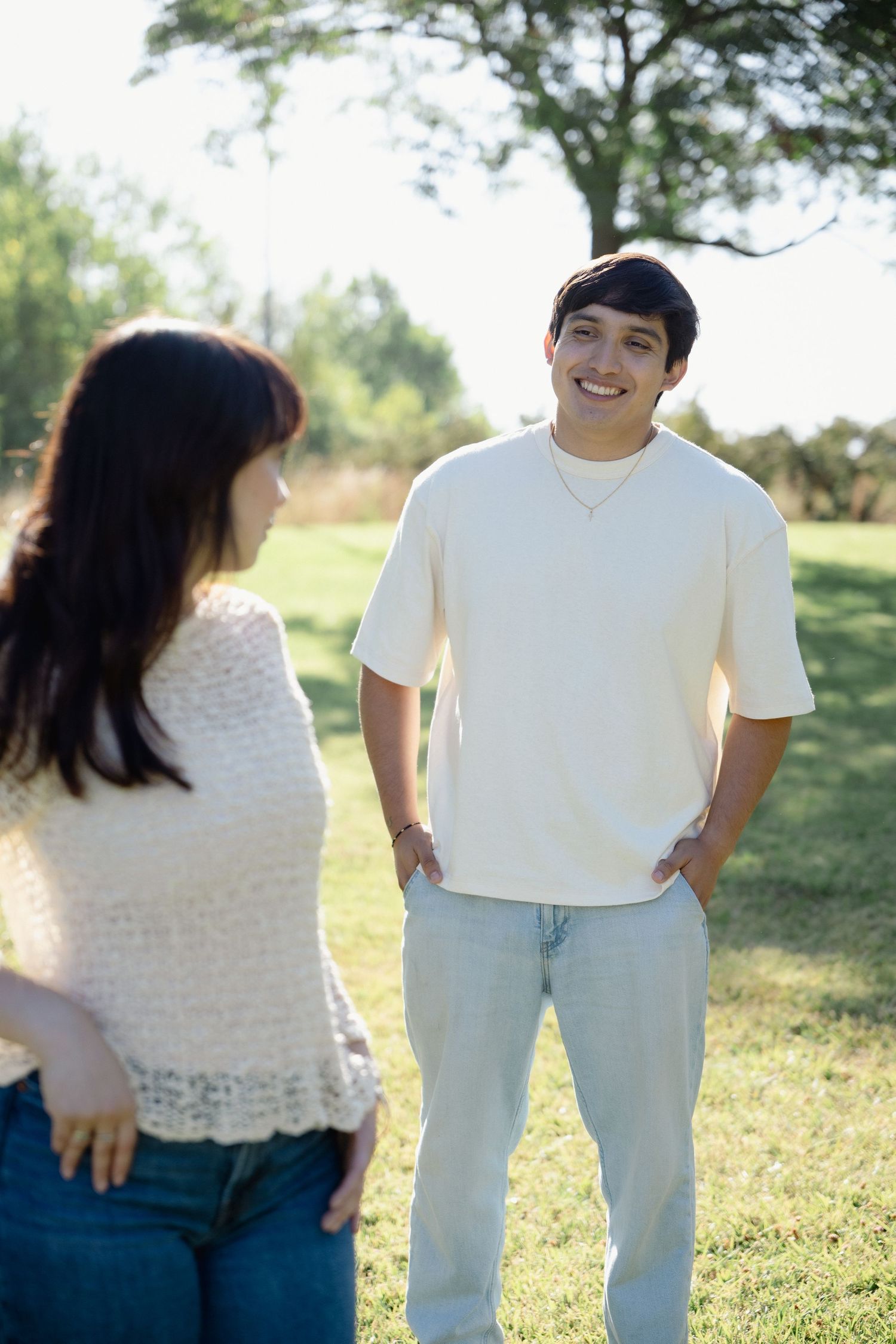 A young man in a cream t-shirt and jeans smiles at a woman in a sunny green park setting.