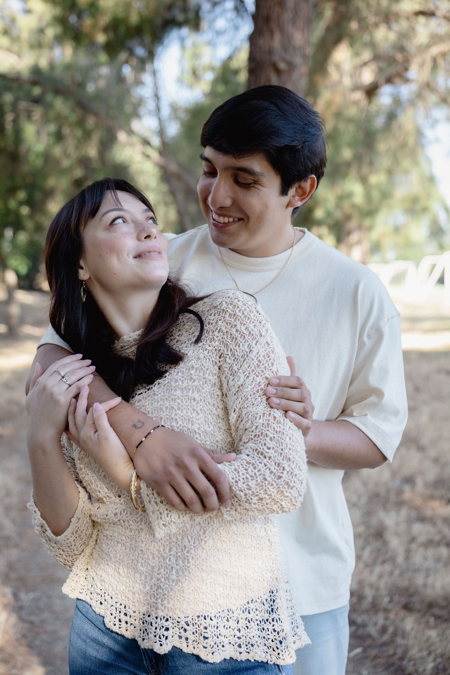 A young couple embraces and smiles at each other outdoors among trees, wearing casual cream and white tops.
