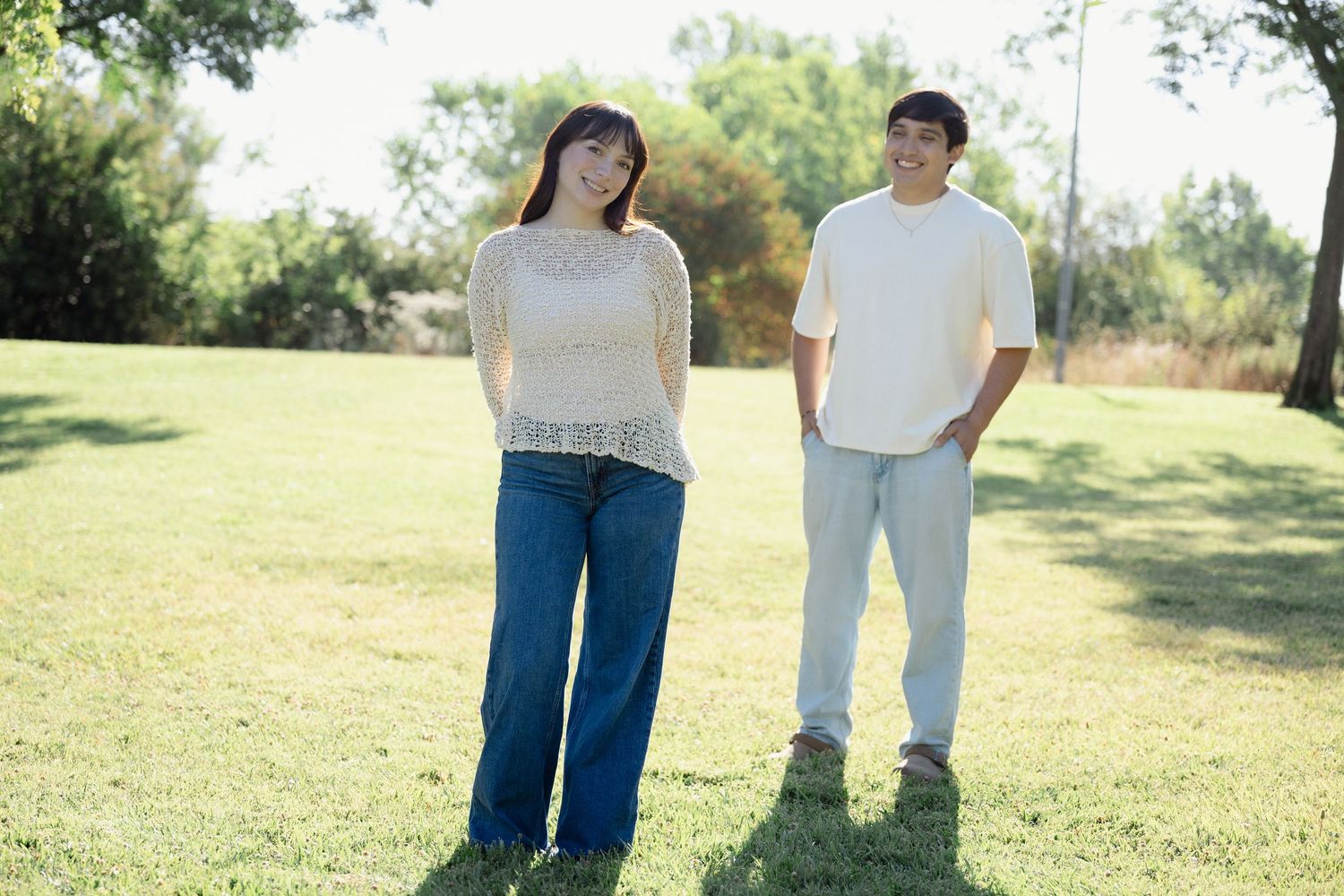 A young Asian couple standing apart on green grass in a sunny park, woman in jeans looking away, man smiling behind her.