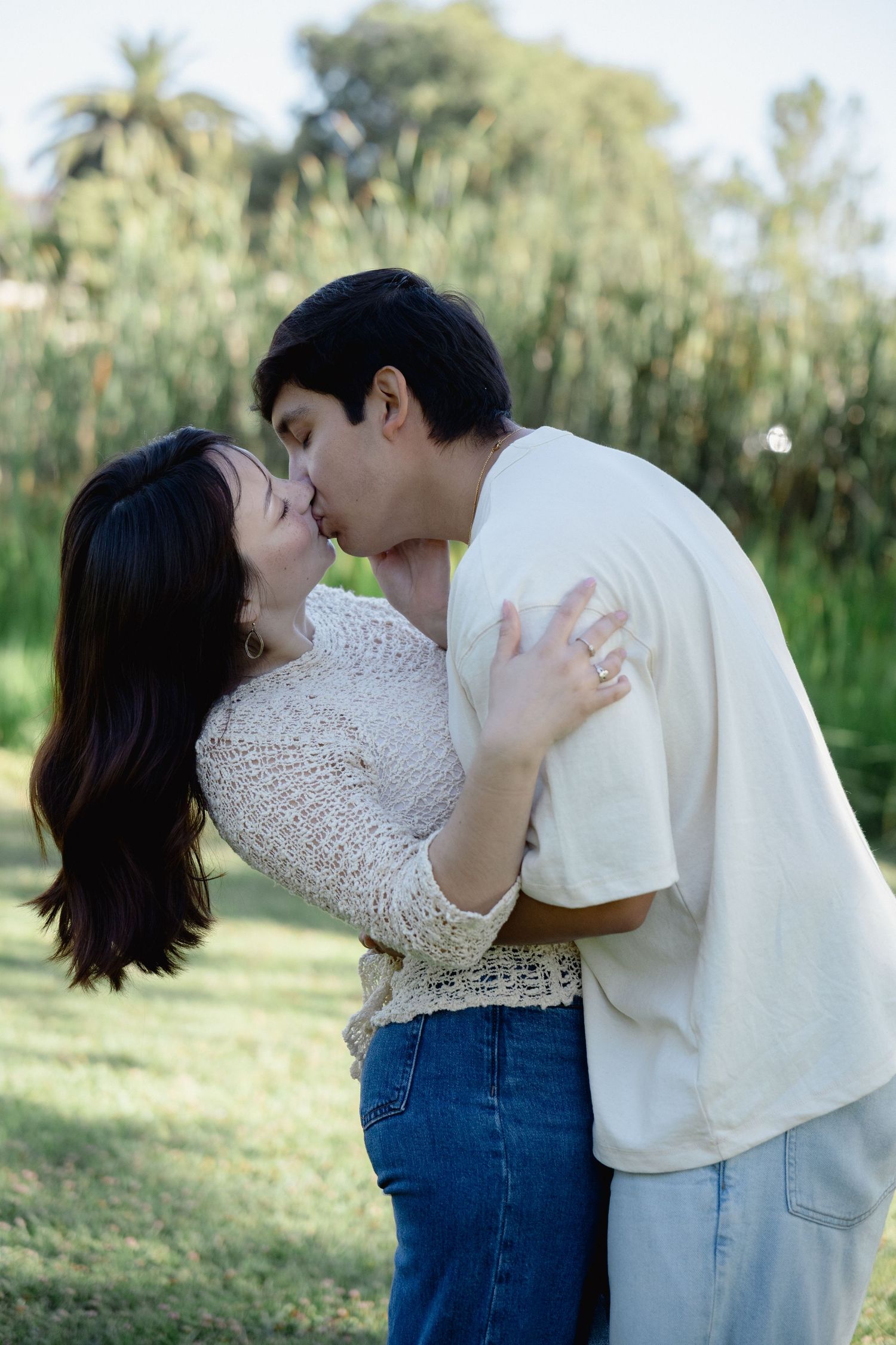A couple shares a romantic kiss outdoors in a lush green park, woman in lace top and jeans, man in white shirt.