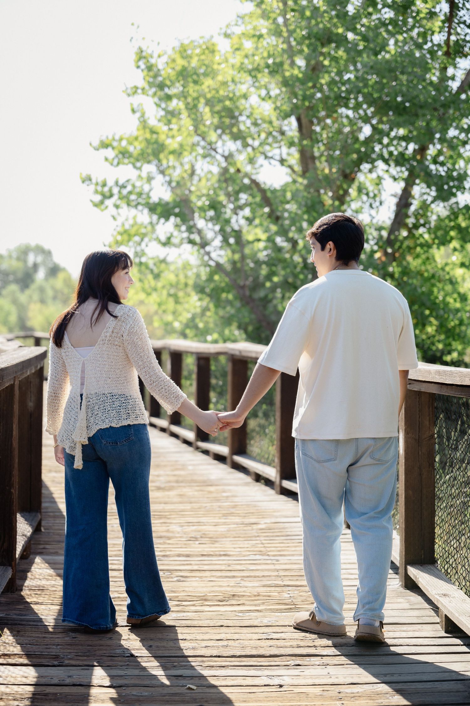 Couple holding hands on a wooden bridge surrounded by lush green trees on a sunny day.