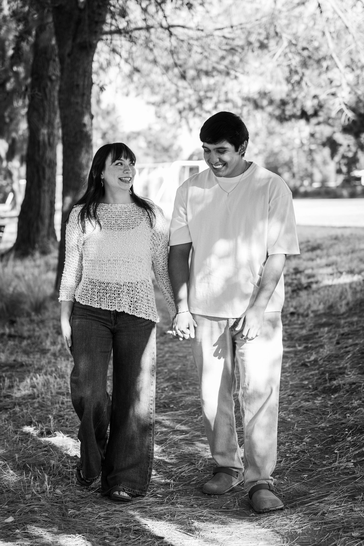 Black and white photo of a happy couple holding hands and walking together along a sunny park path.