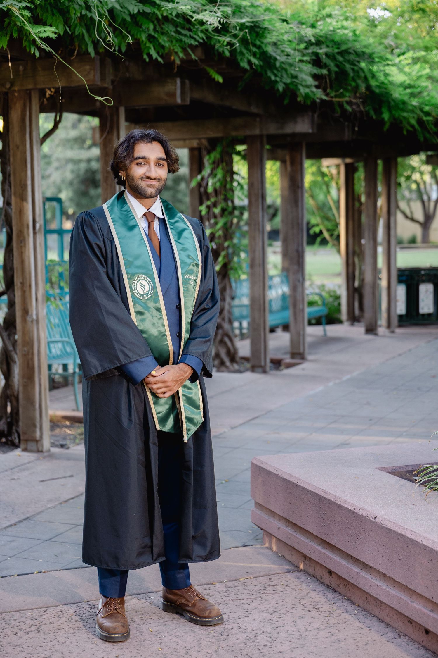Young man in black graduation gown and green stole smiling outdoors near wooden pergola with lush greenery.