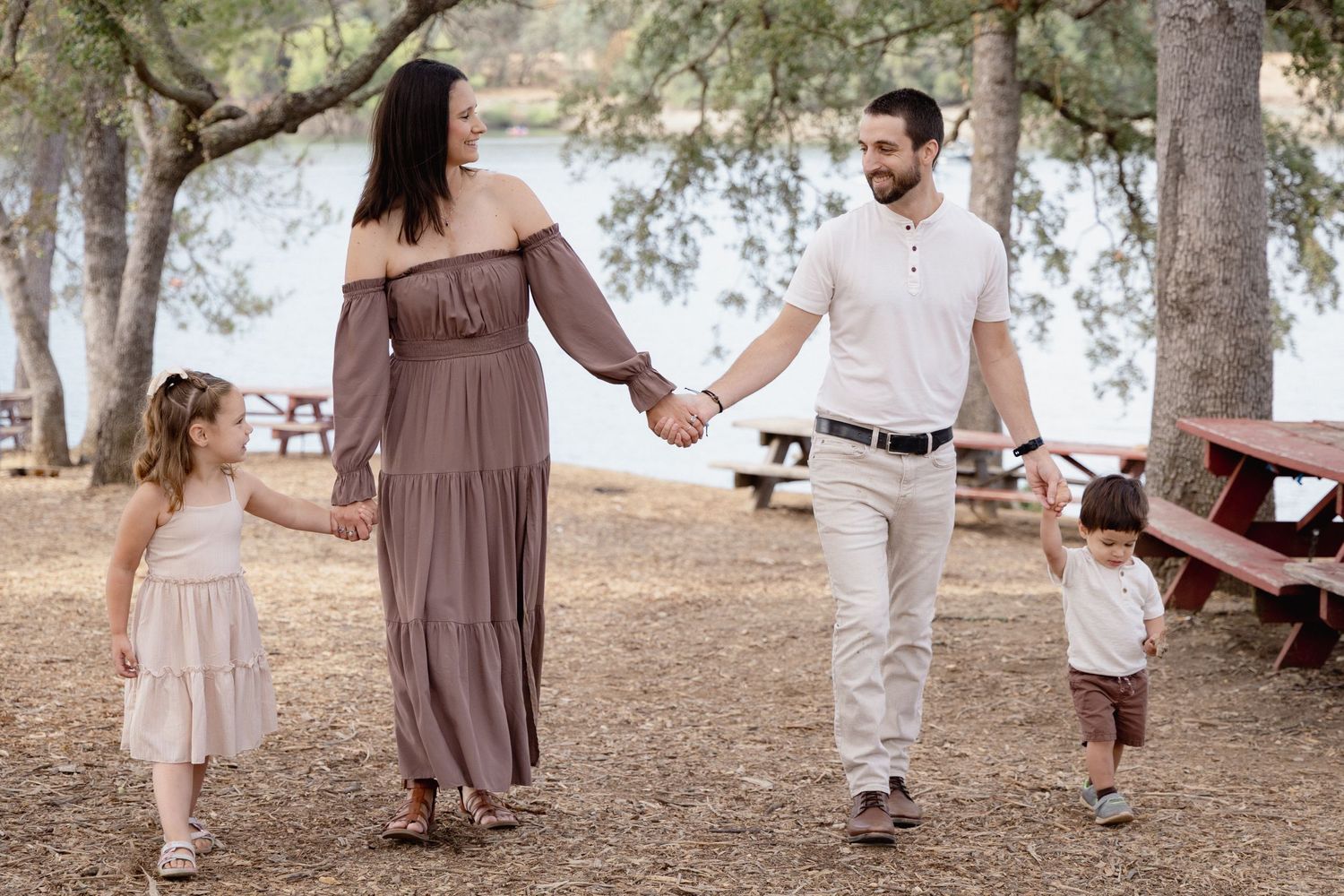 Family of four holding hands and walking together in a scenic park near a lake on a sunny day.